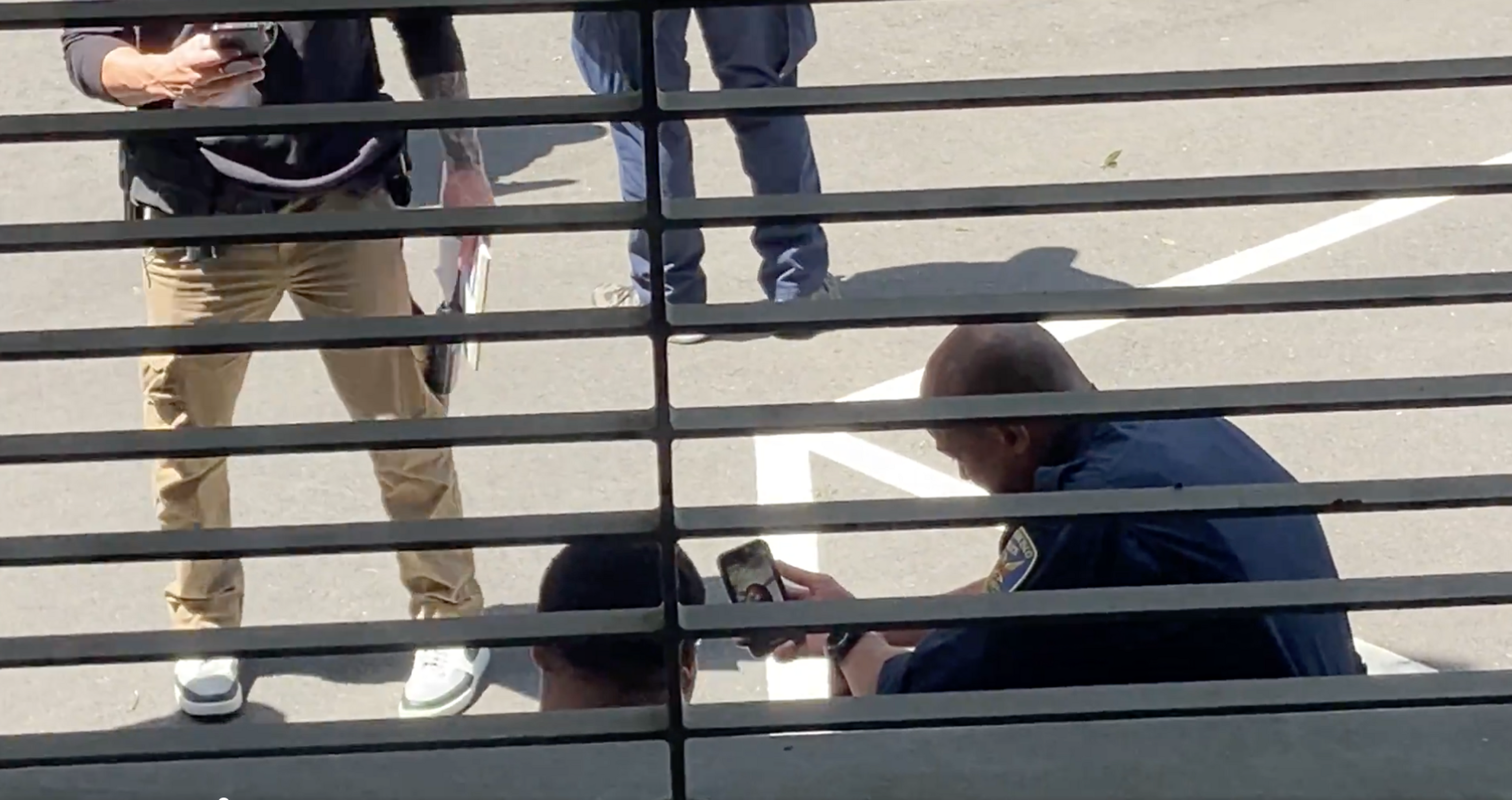 Three people stand behind horizontal bars; one in a blue uniform is kneeling, looking at a phone.