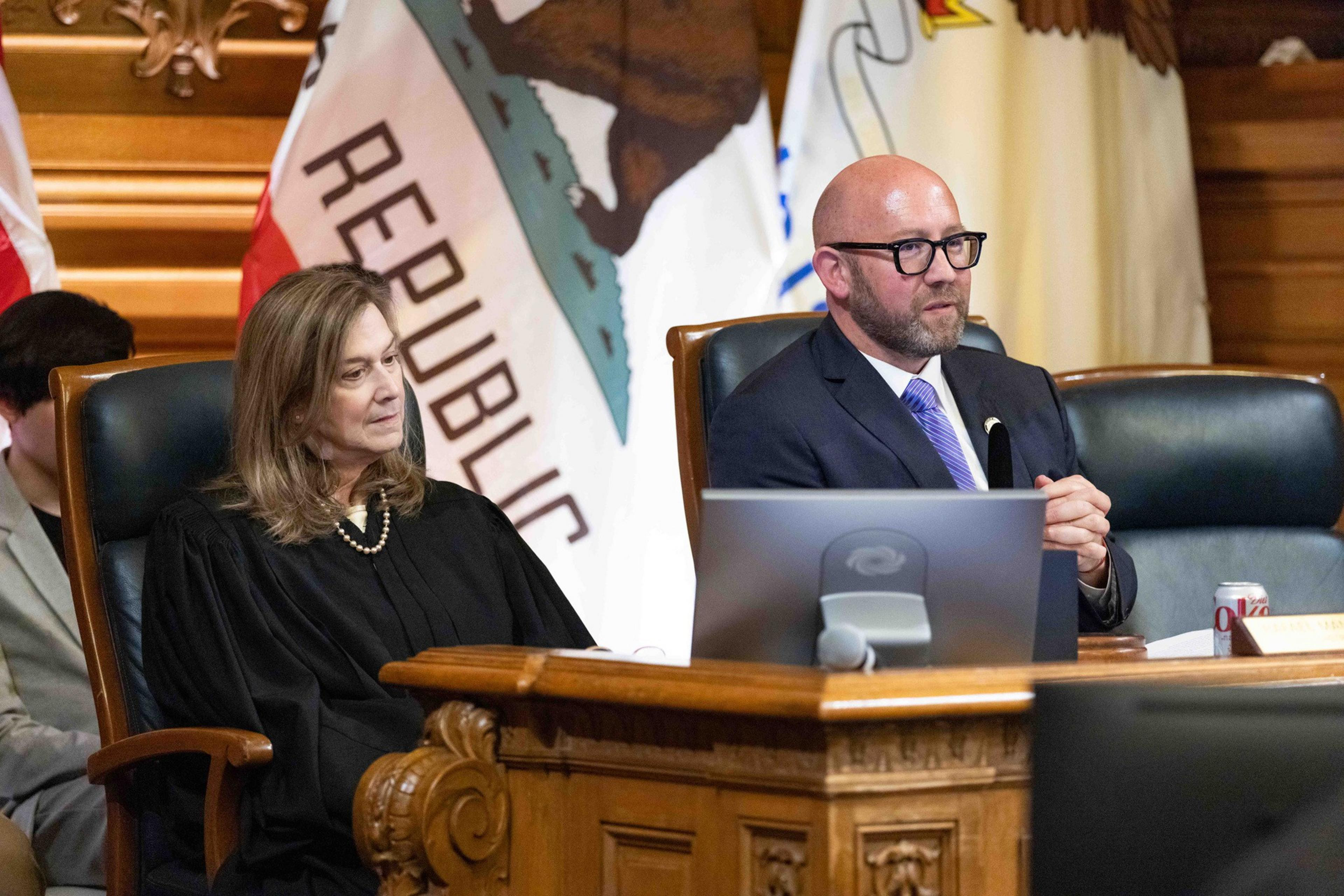 A woman in a black robe sits beside a man in a suit at a wooden podium, with flags in the background. The scene appears formal and official.
