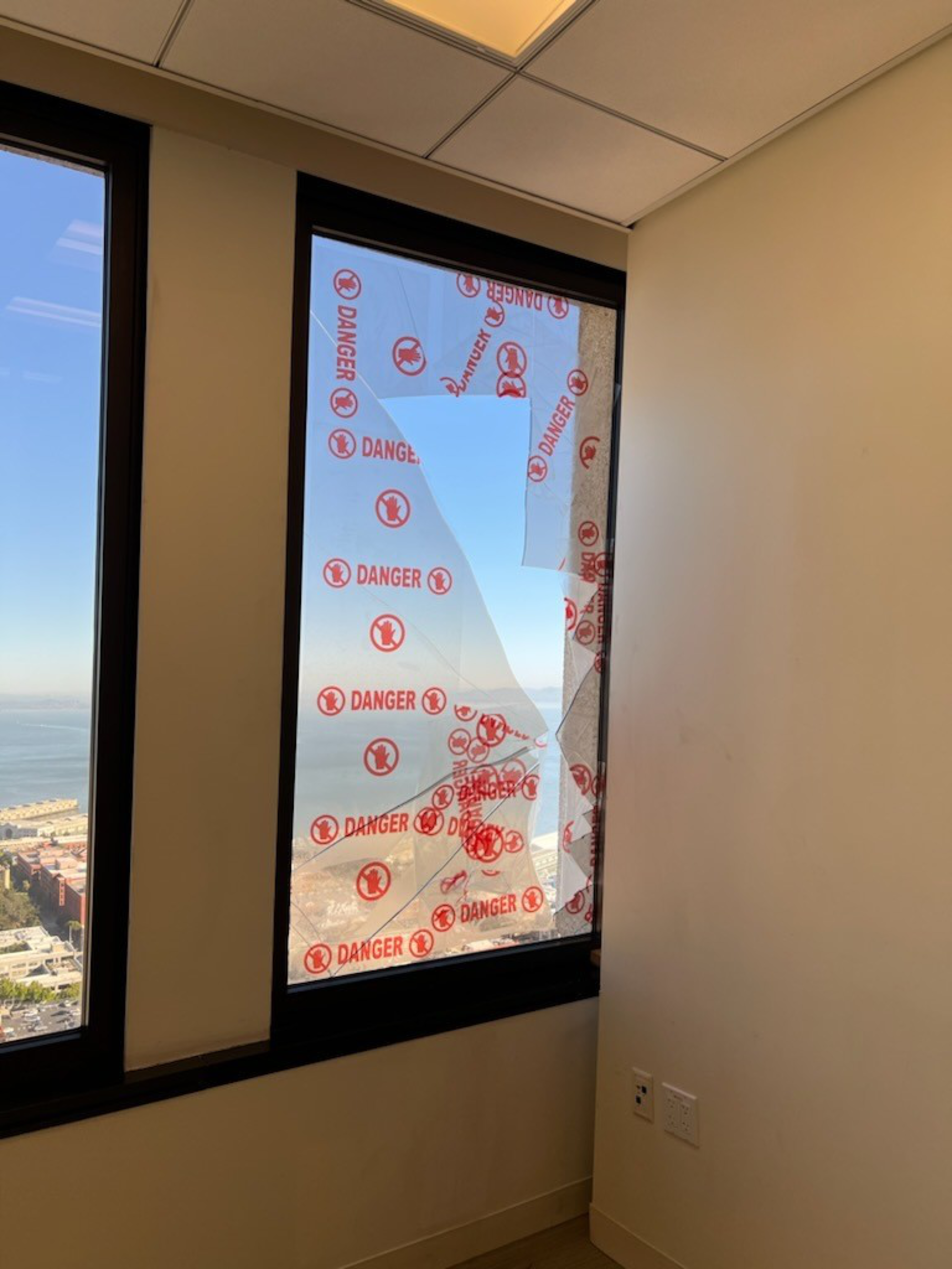 A room with a broken window covered in red &quot;DANGER&quot; tape, showing a high-altitude cityscape and waterfront outside.