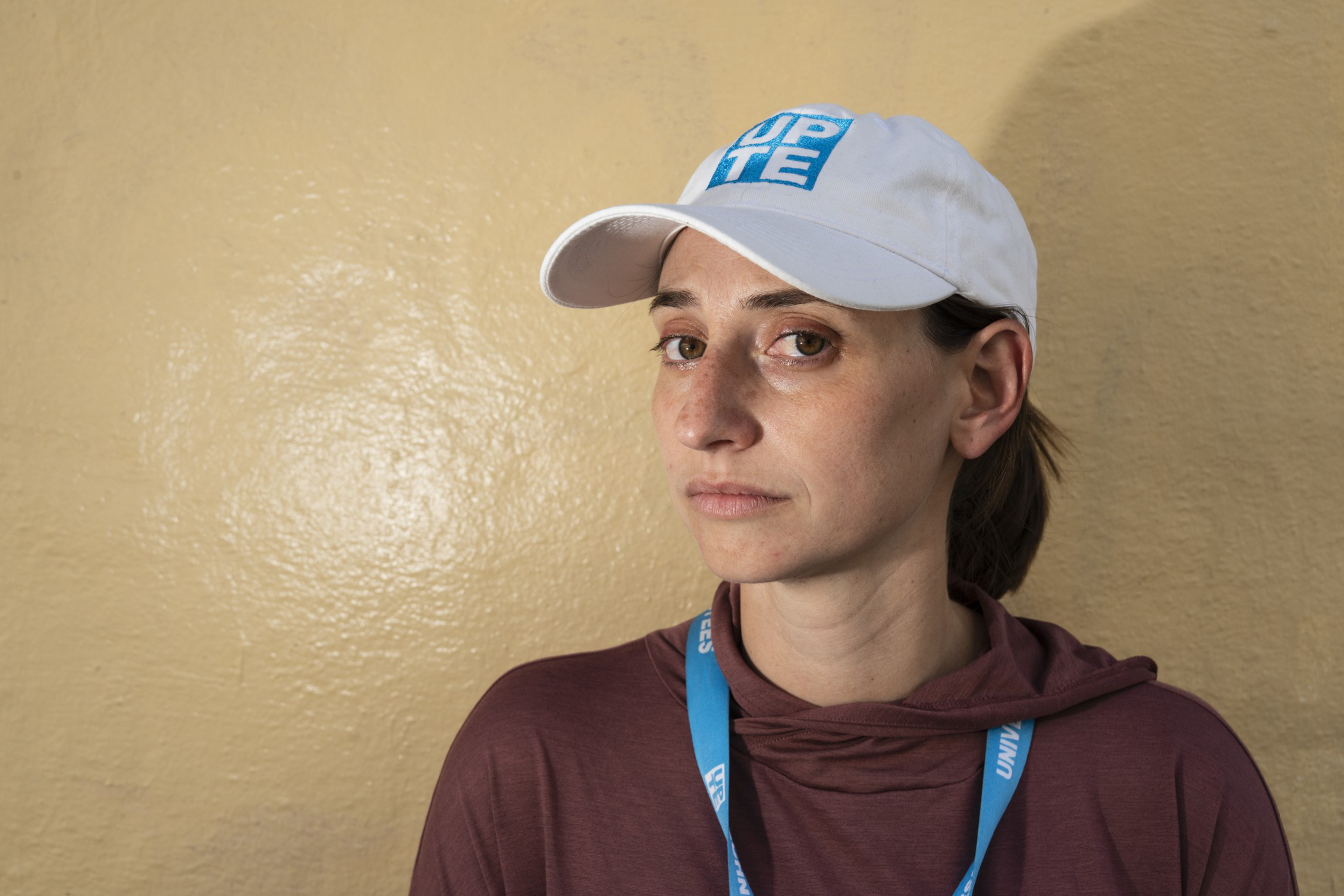 A woman wearing a white cap with blue writing, a maroon hoodie, and a blue lanyard looks slightly to the side against a beige wall.