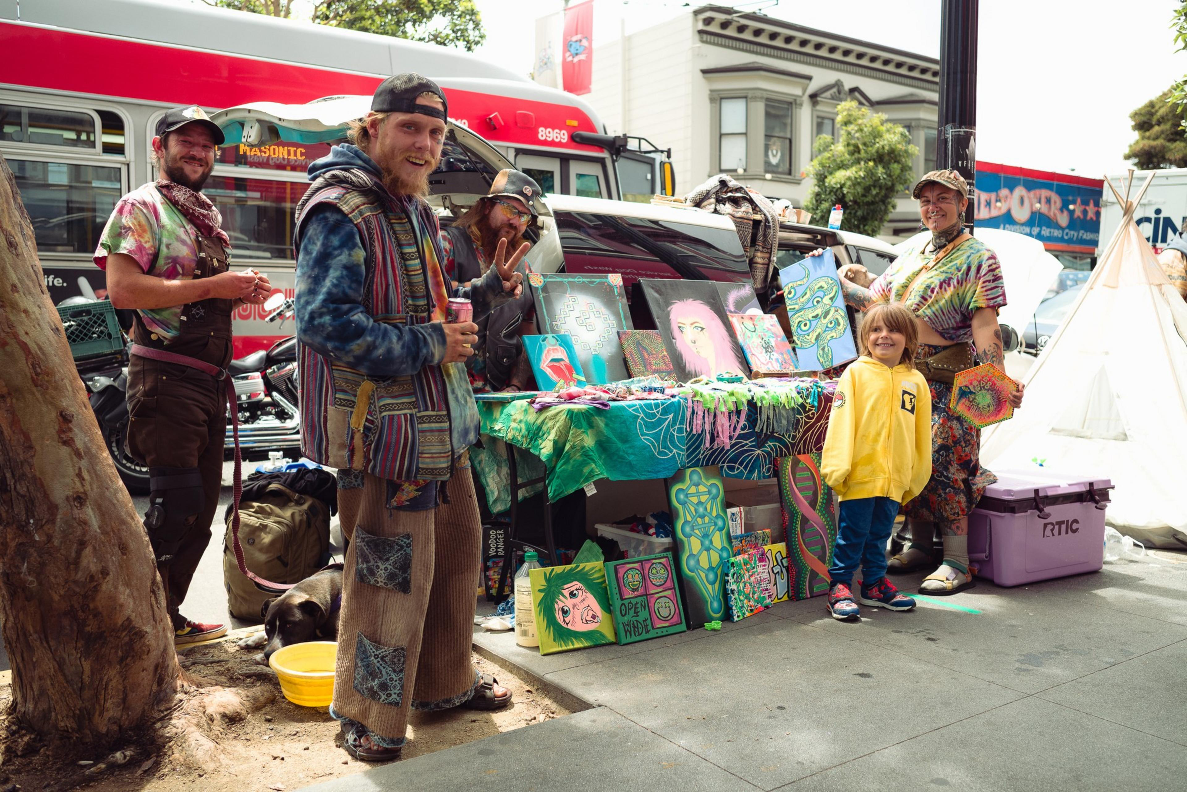 Four adults and one child stand by a colorful art display on a busy sidewalk with a red bus and buildings in the background.