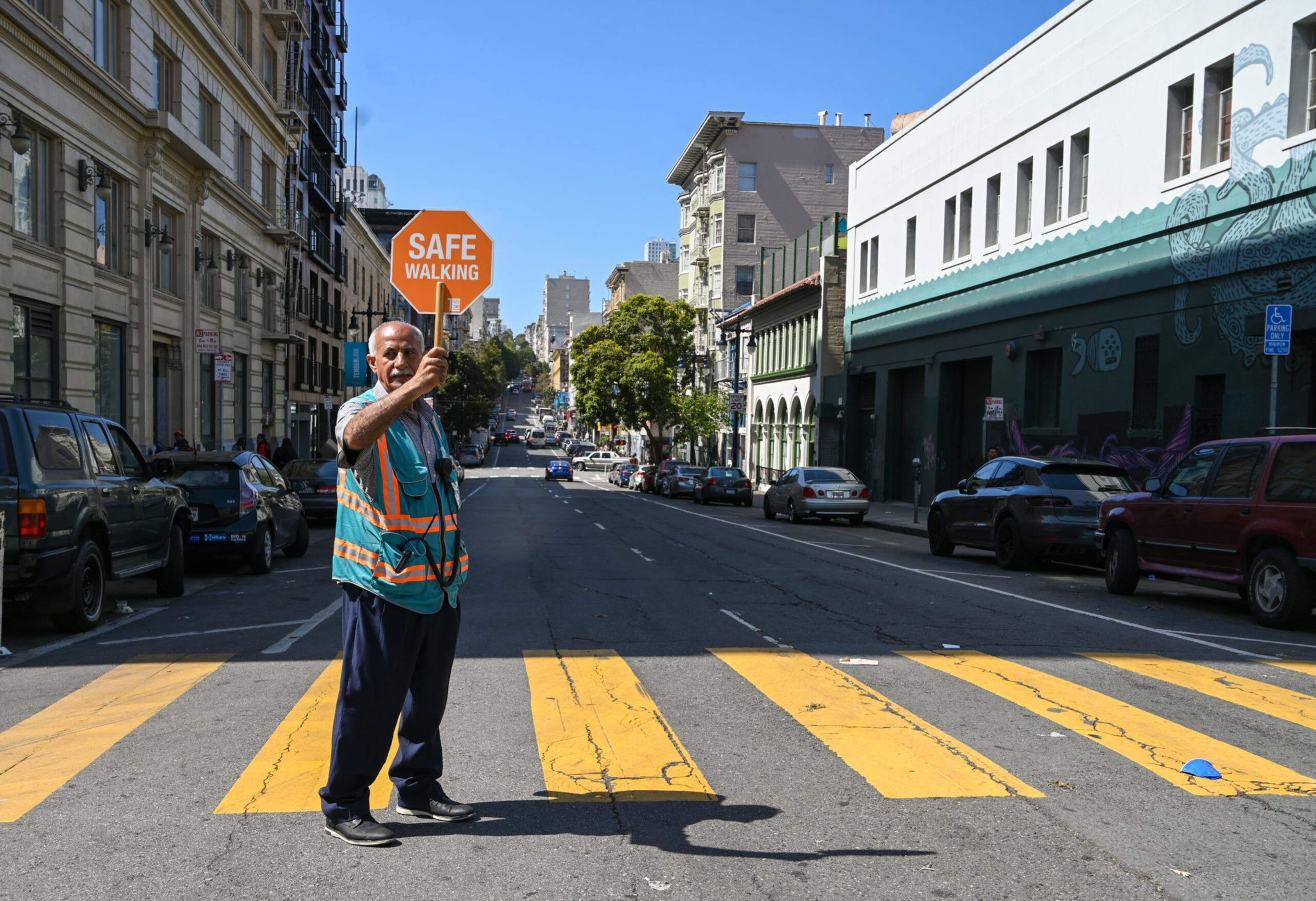 A person in a hi-vis vest holds a &quot;SAFE WALKING&quot; sign on a sunlit, crosswalk-lined street with cars and buildings.