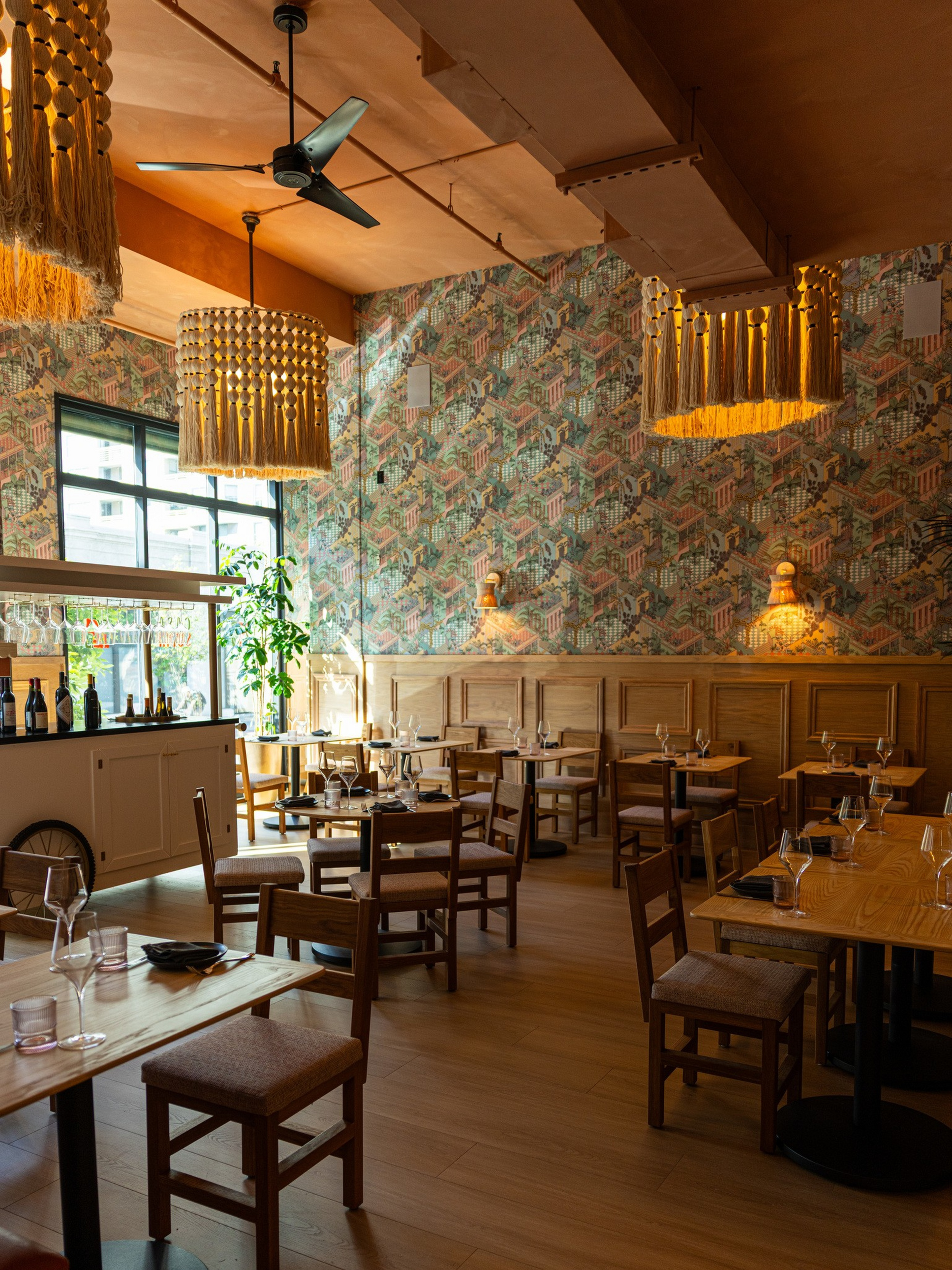Wood-toned dining tables and chairs fill a warmly lit restaurant with patterned wallpaper, large macramé pendant lights, and a ceiling fan by tall windows.