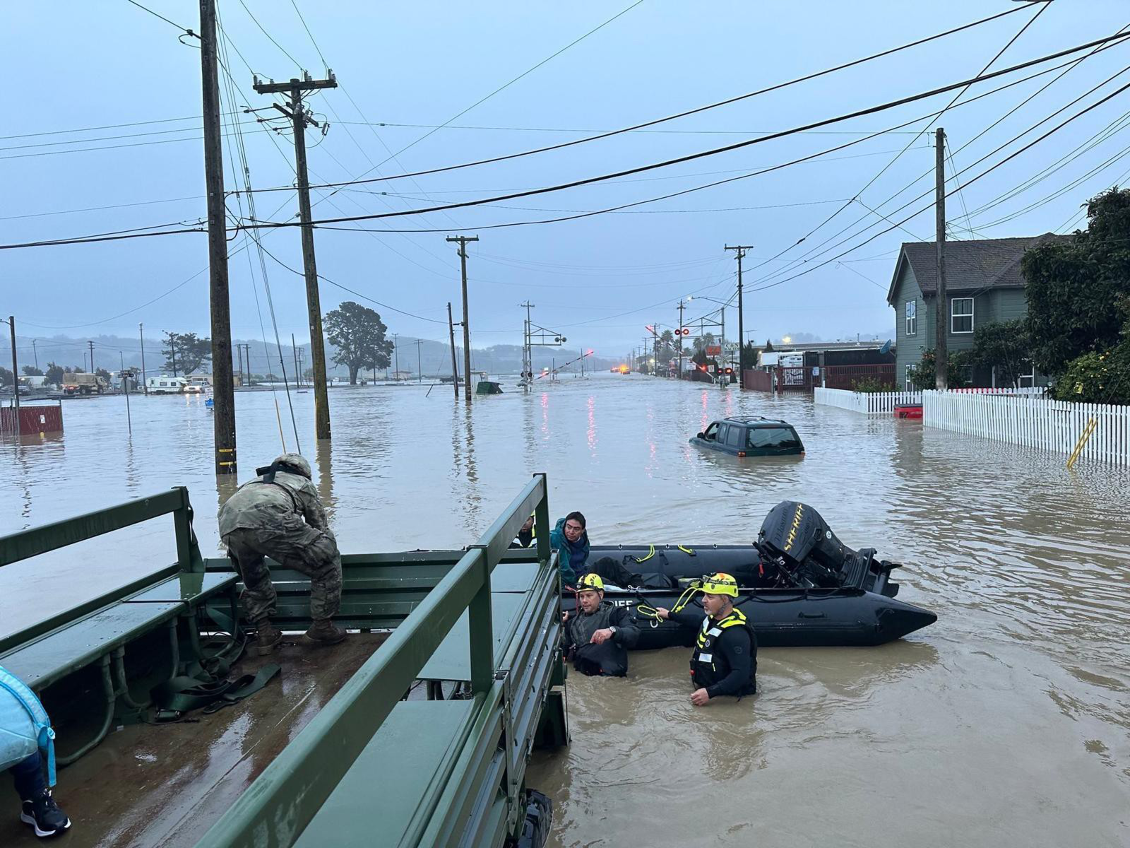 Floodwaters cover a street with a partially submerged car, while rescue workers in wetsuits assist people into a boat beside a military truck.