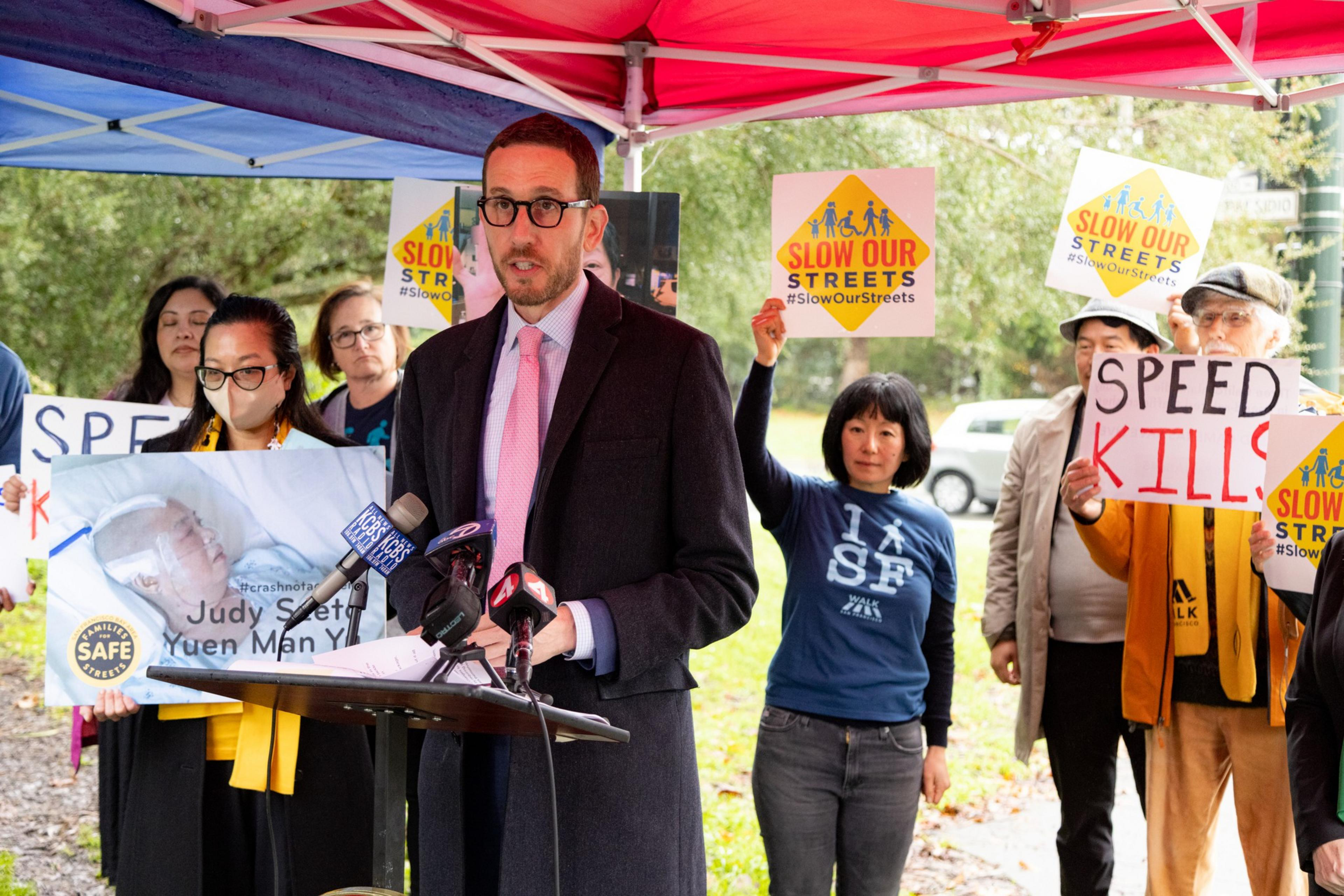 A man in a suit stands in front of a podium backed by people holding signs that say "speed kills".