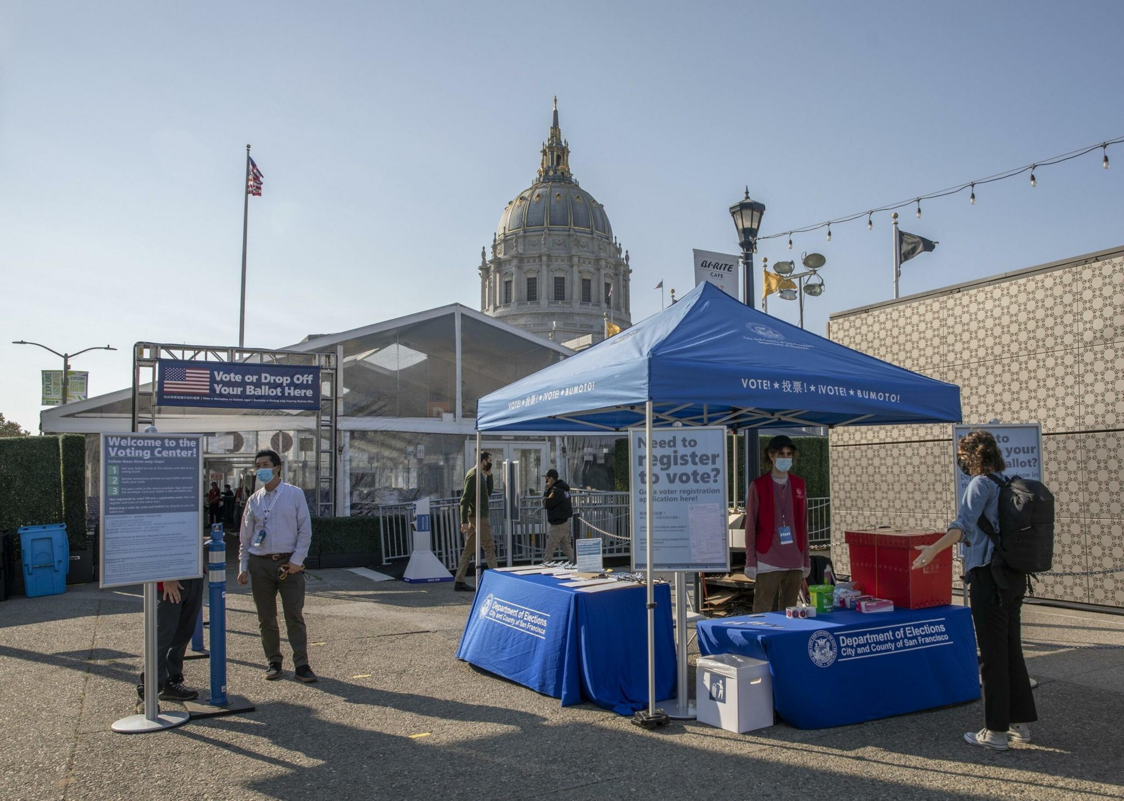 A blue tent with voting signs and people wearing masks outside a grandiose building.