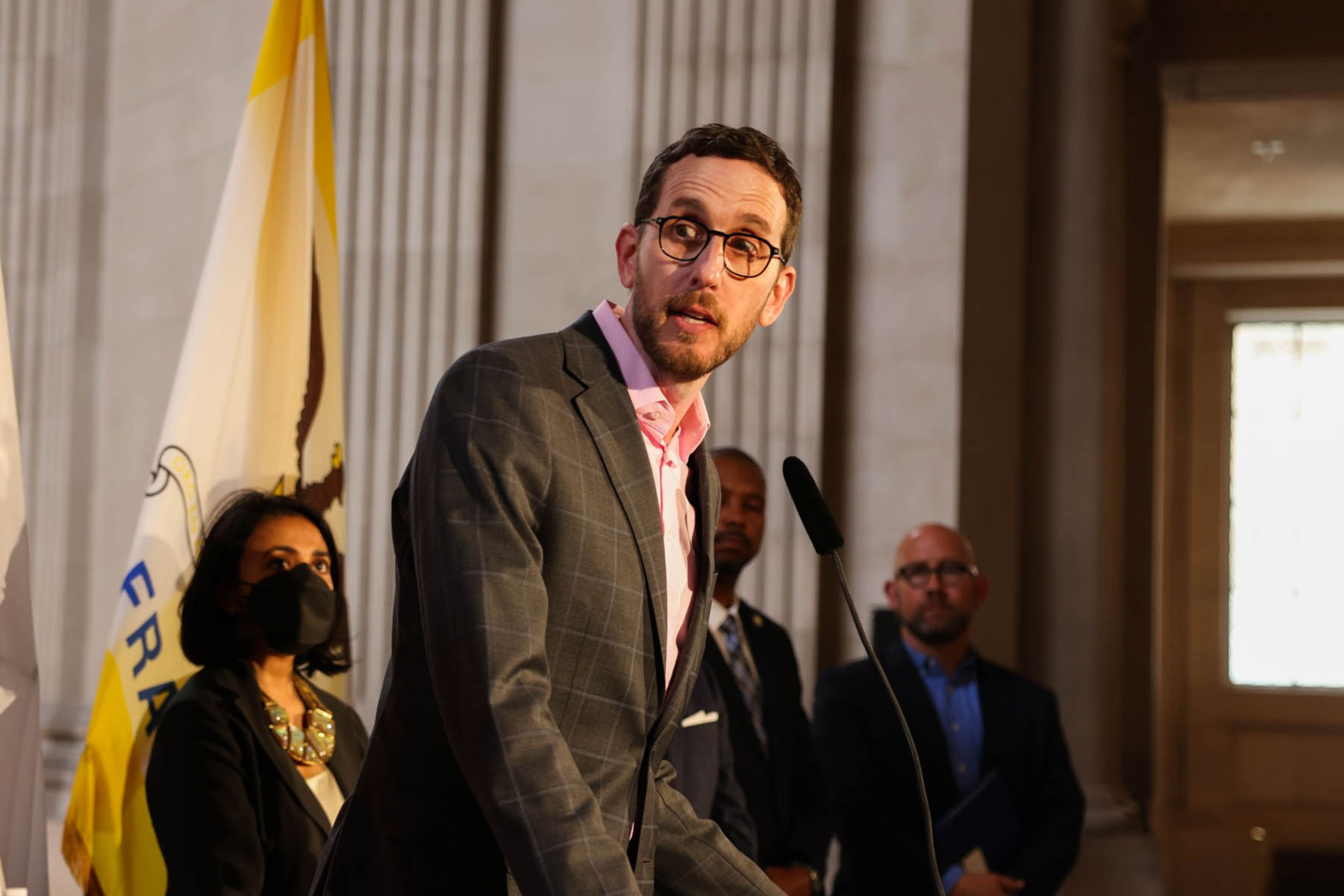 A man in glasses and a gray suit speaks at a podium while three people stand behind him inside a formal building.
