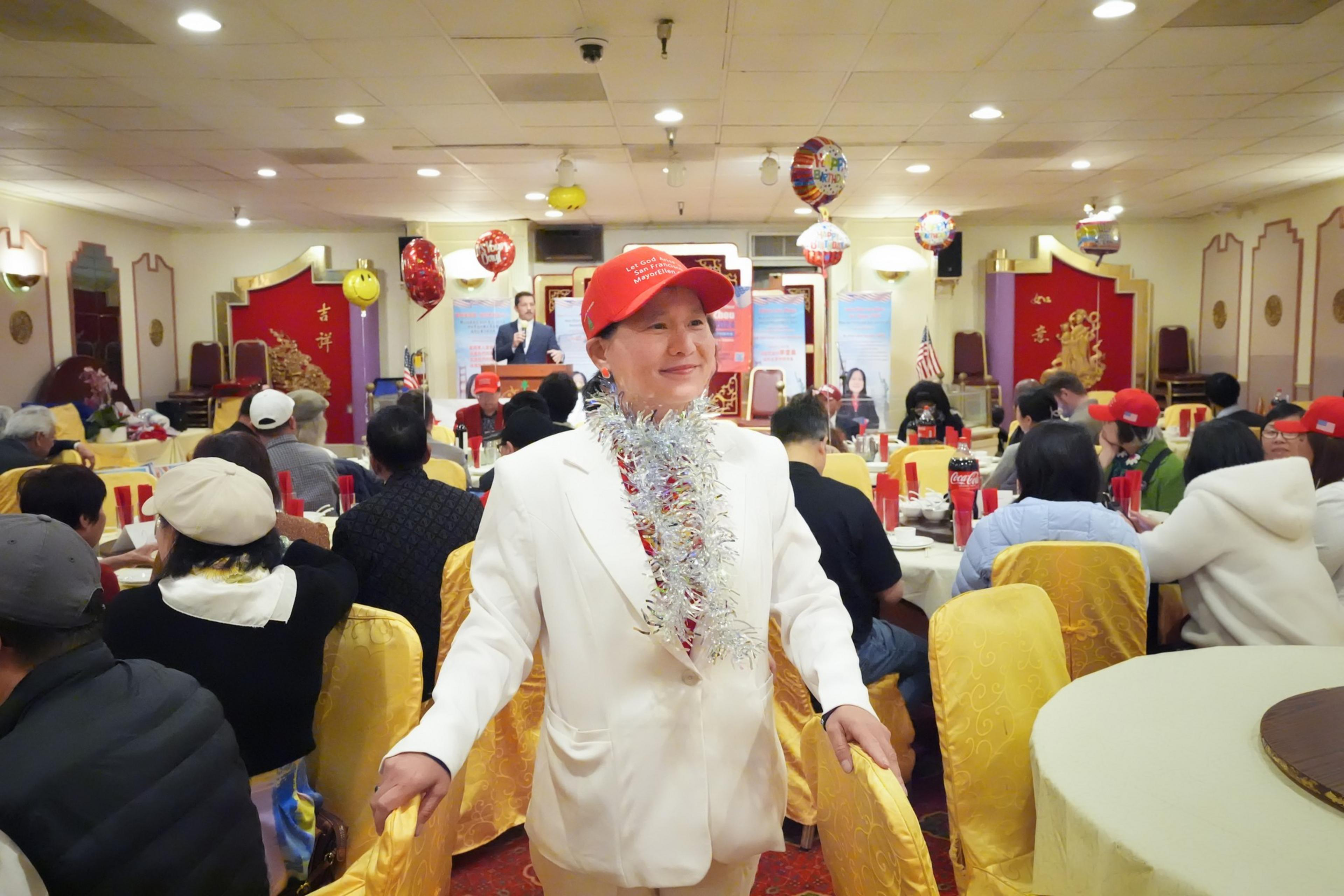 A person in a white suit and red cap stands in a festive room full of people wearing similar caps, facing a stage with a speaker.