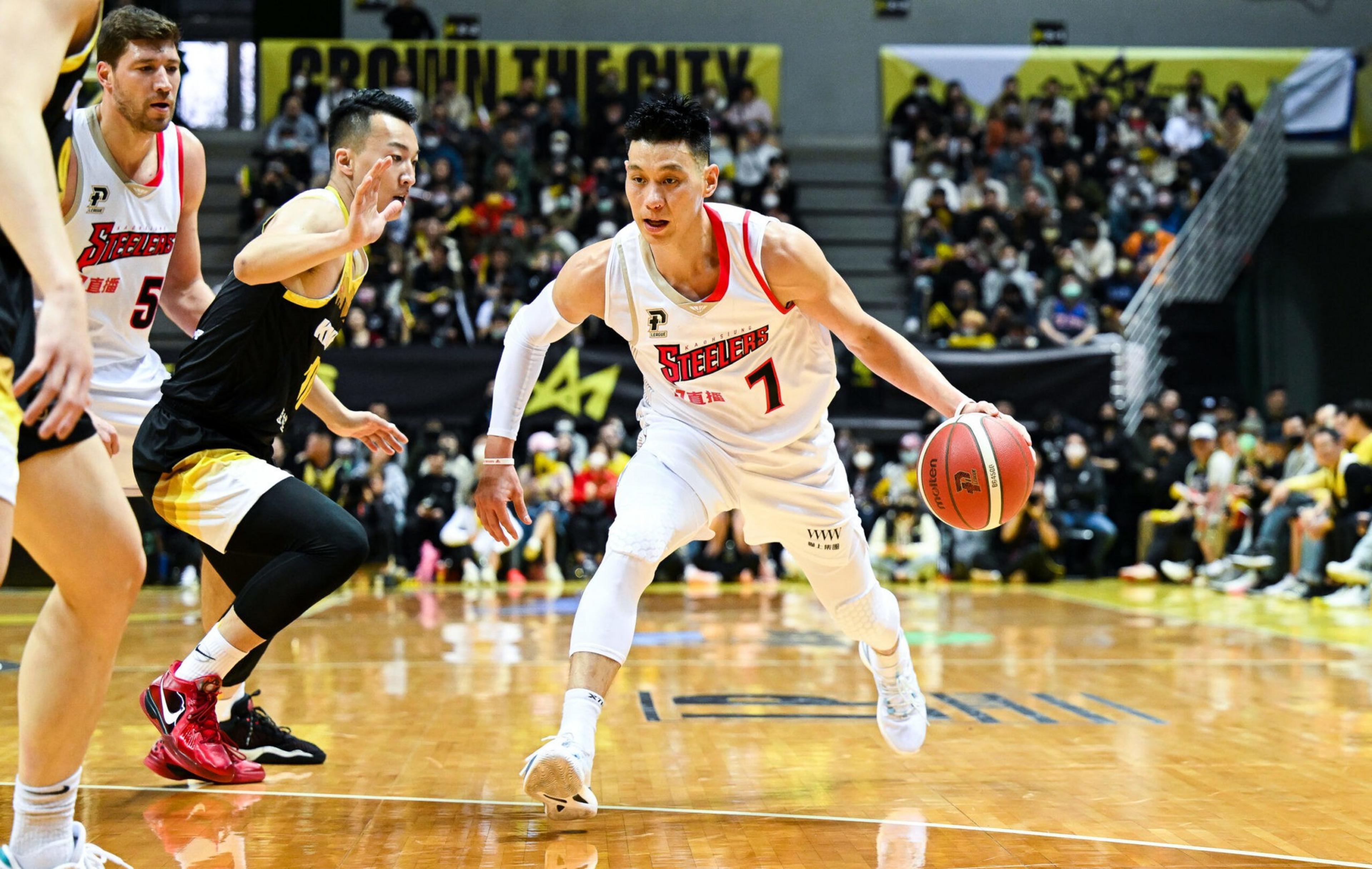 A basketball player wearing a white Steelers jersey dribbles the ball while being closely defended by a player in a black and yellow uniform on a court with a crowded audience.