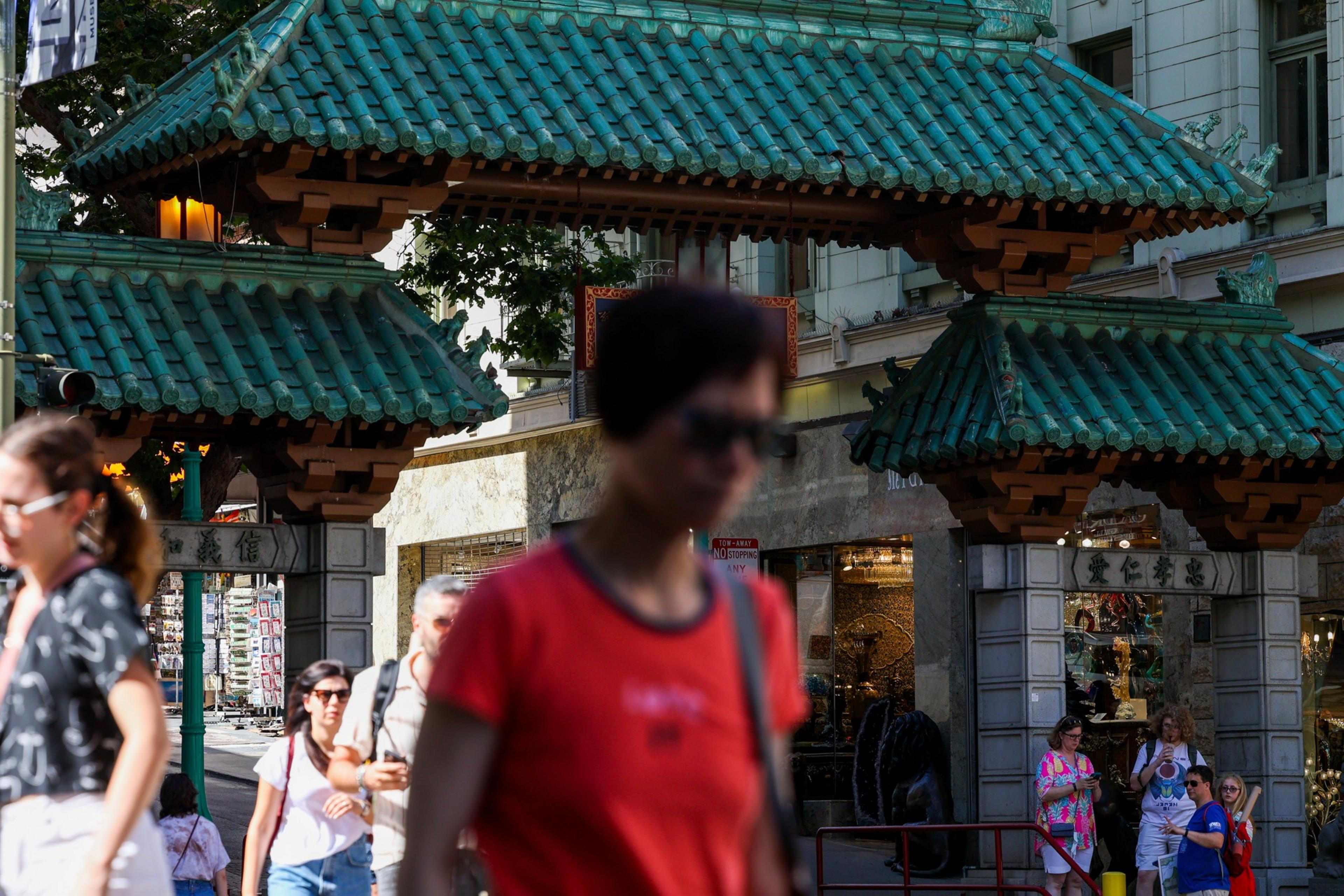 A green-tiled traditional Chinese-style gate stands in a busy urban area with several people walking or standing around it.