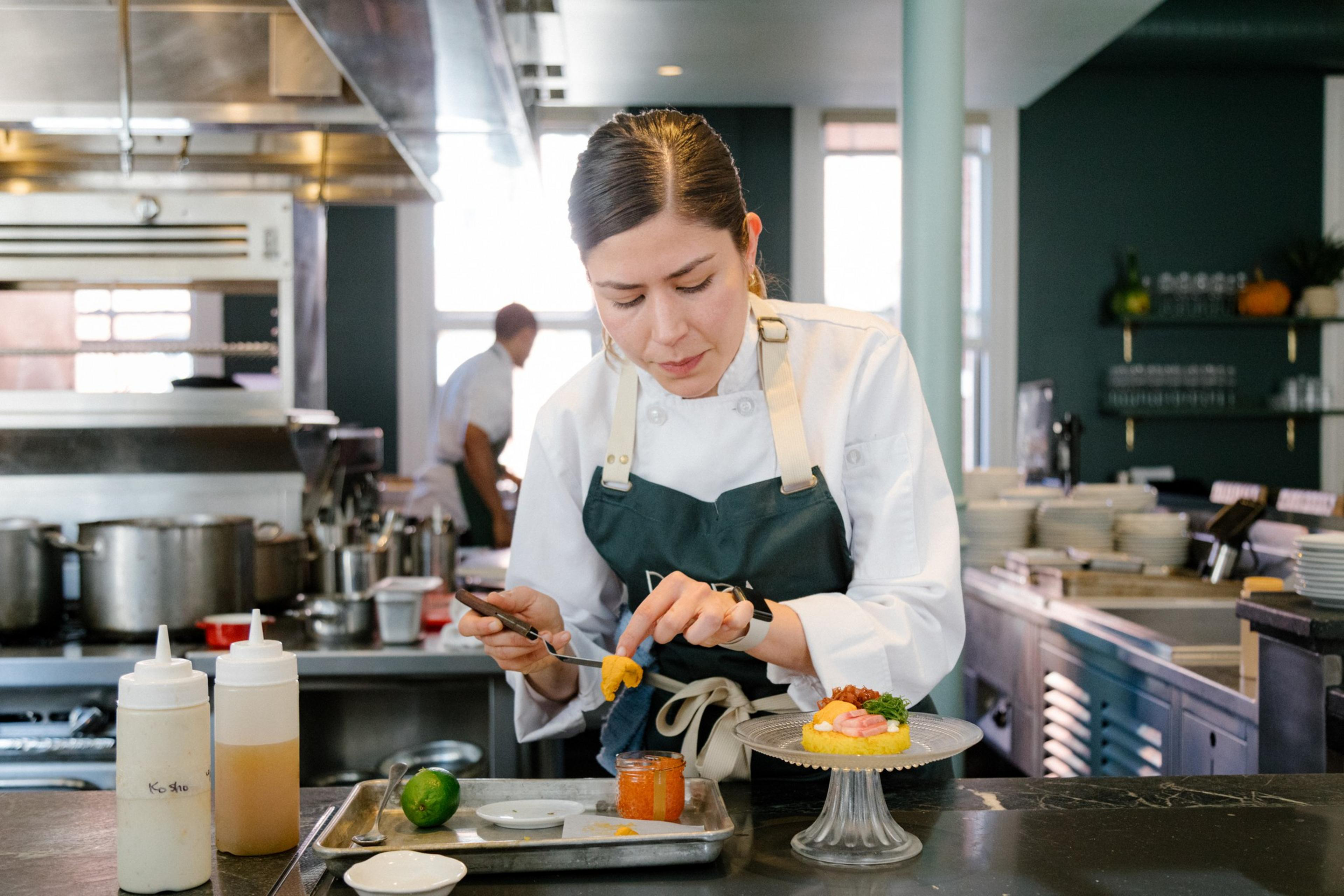 A chef in a white coat and dark apron is garnishing a dish with an orange ingredient in a professional kitchen, with pots and ingredients visible around.