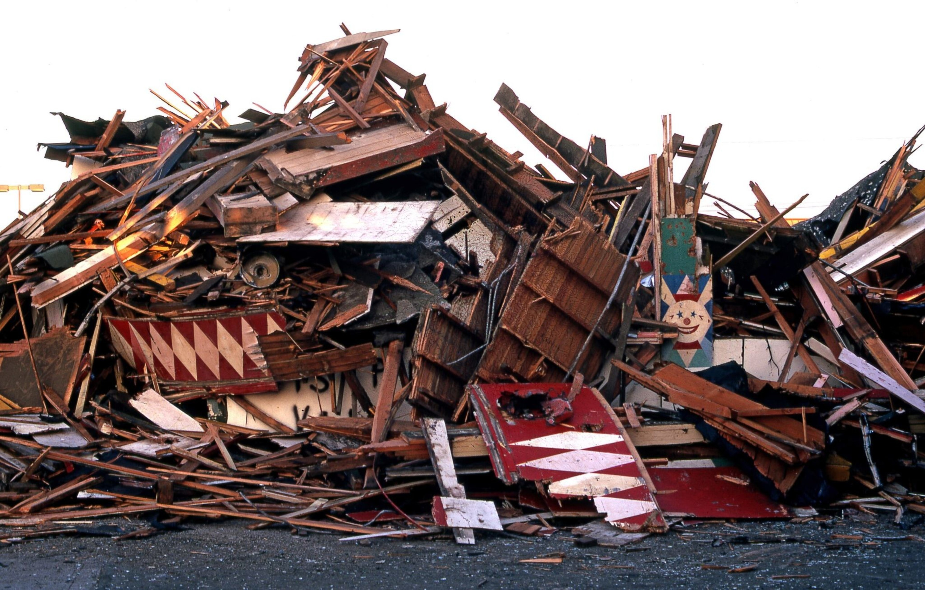 A large pile of broken wooden boards and debris, some painted with red and white patterns and a smiling clown face on one plank.