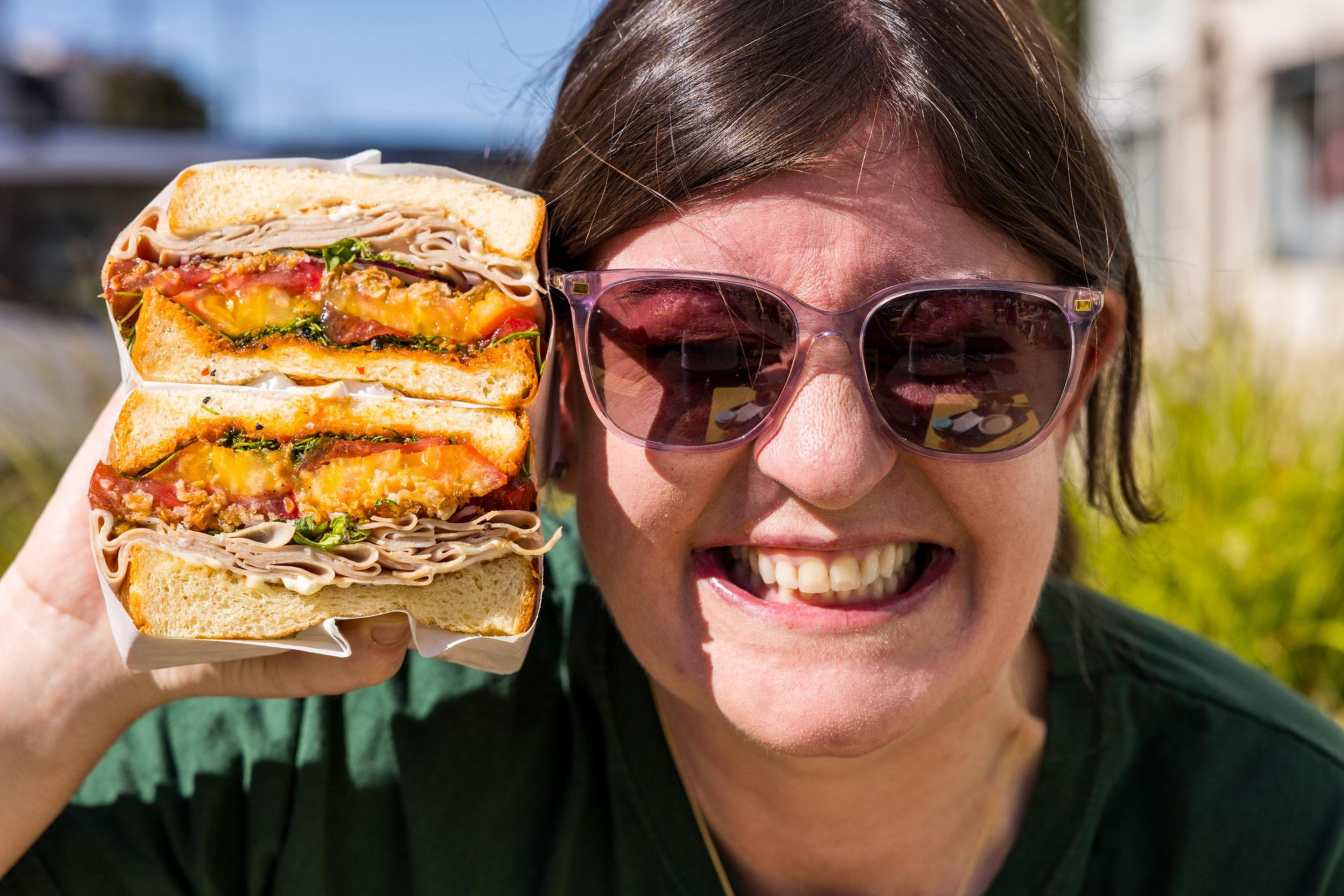 A person wearing sunglasses smiles widely, holding a large sandwich filled with turkey, melted cheese, bacon, spinach, and tomato close to their face.