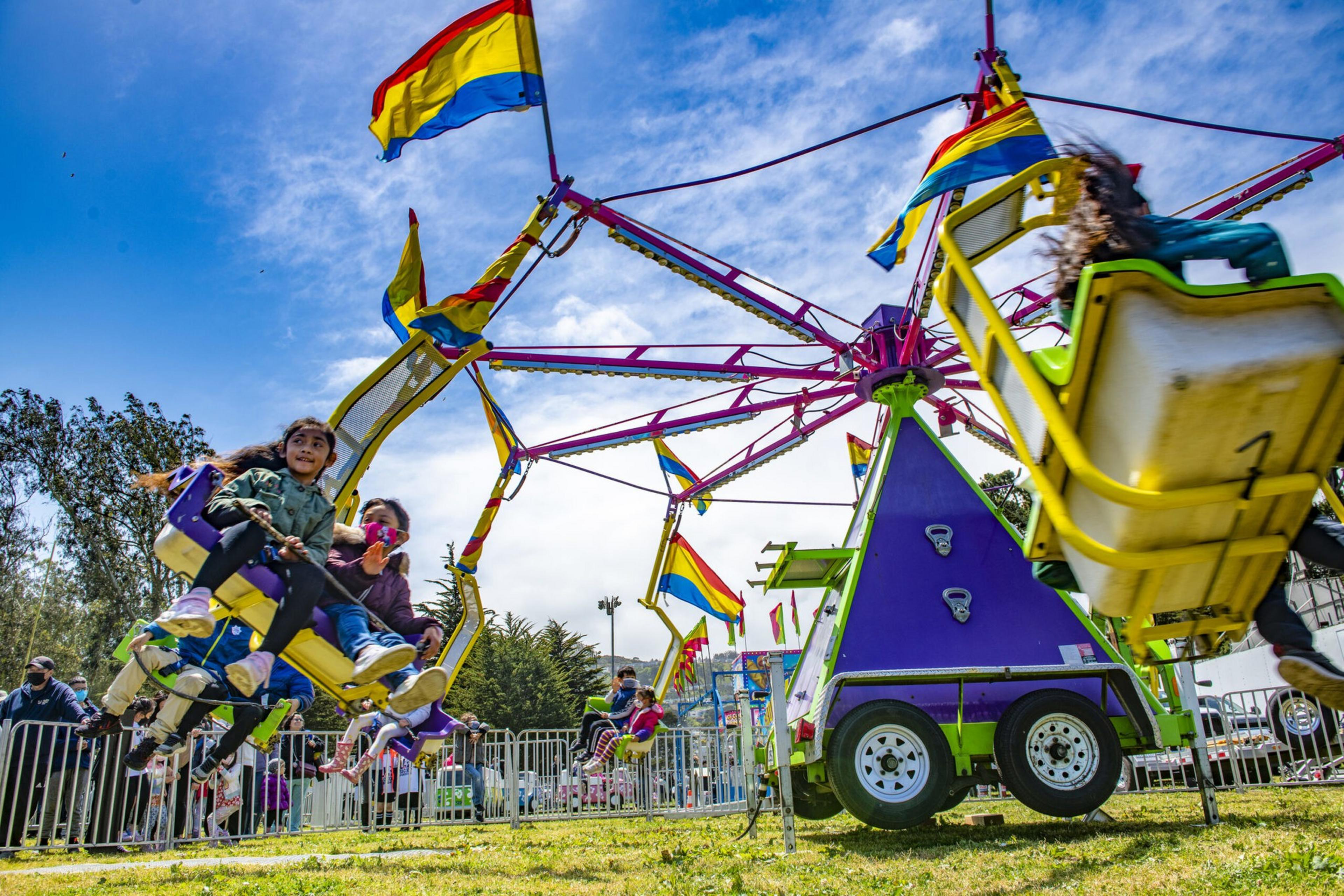 Children are enjoying a colorful swing ride with red, yellow, and blue flags on a sunny day at an outdoor fair.