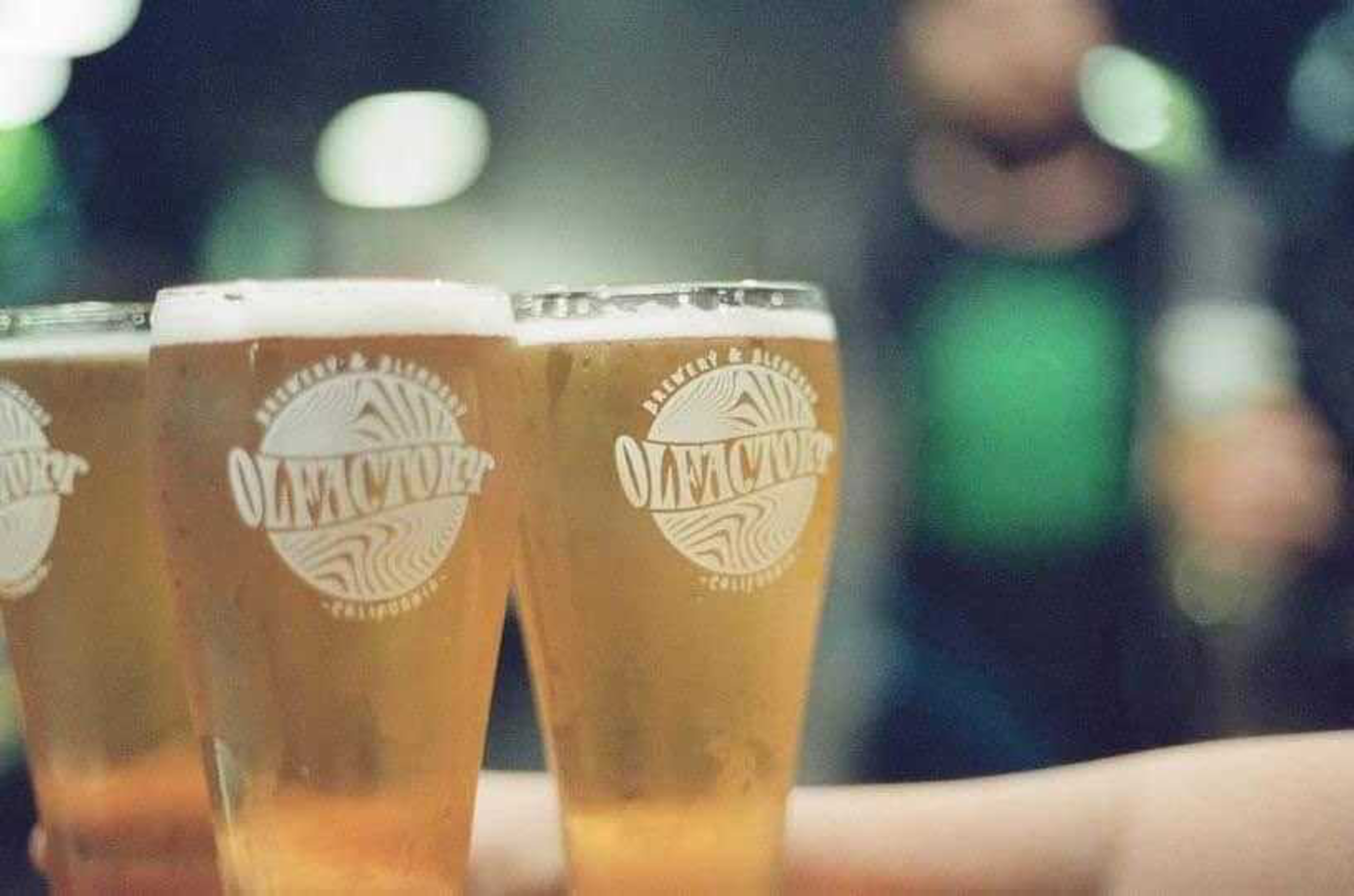 Three glasses filled with light amber beer, each branded with a circular logo, are held up together with blurred people in the background.