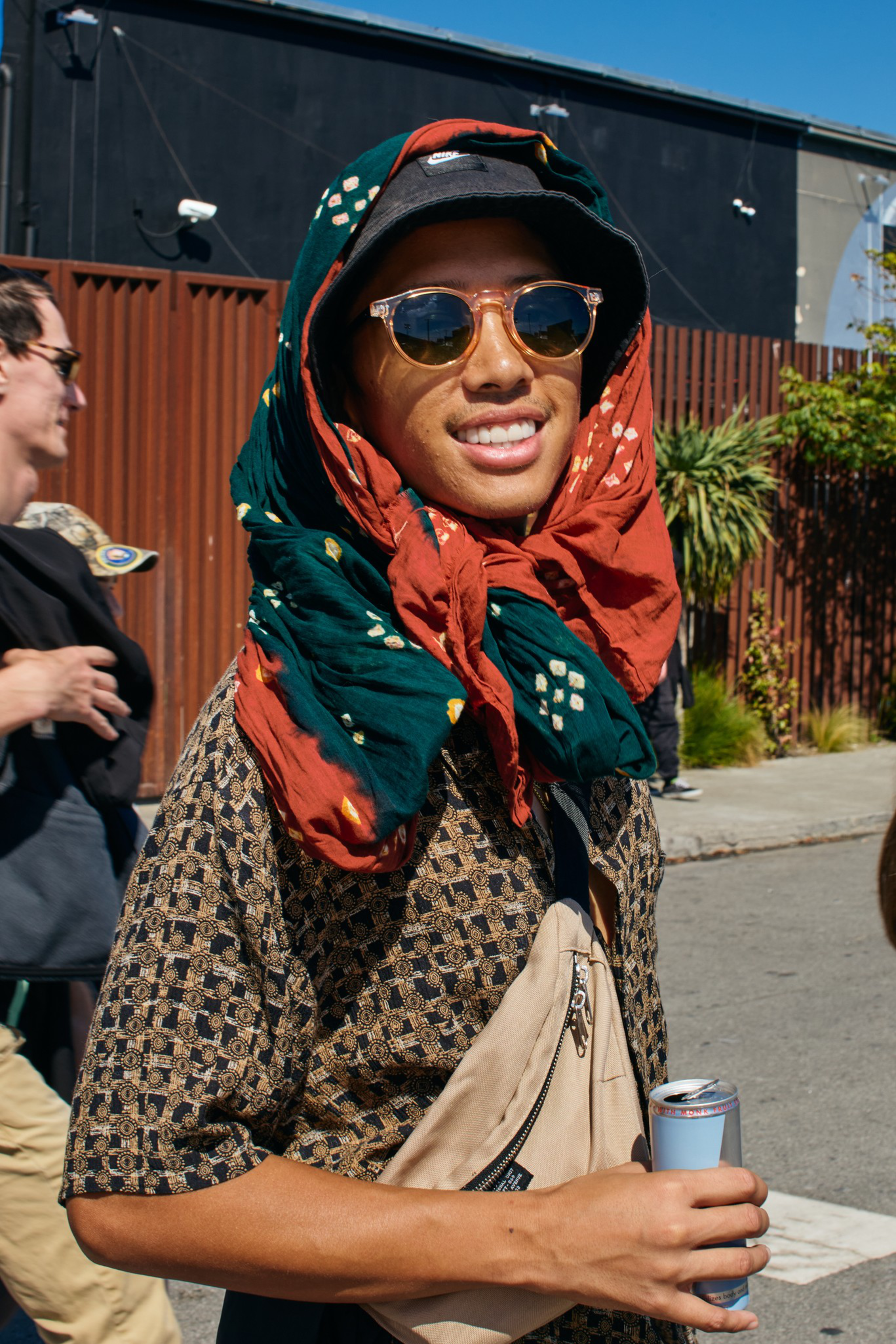 A smiling person wears round sunglasses, a patterned shirt, a multi-colored scarf over a black hat, and holds a can while standing outdoors on a sunny day.