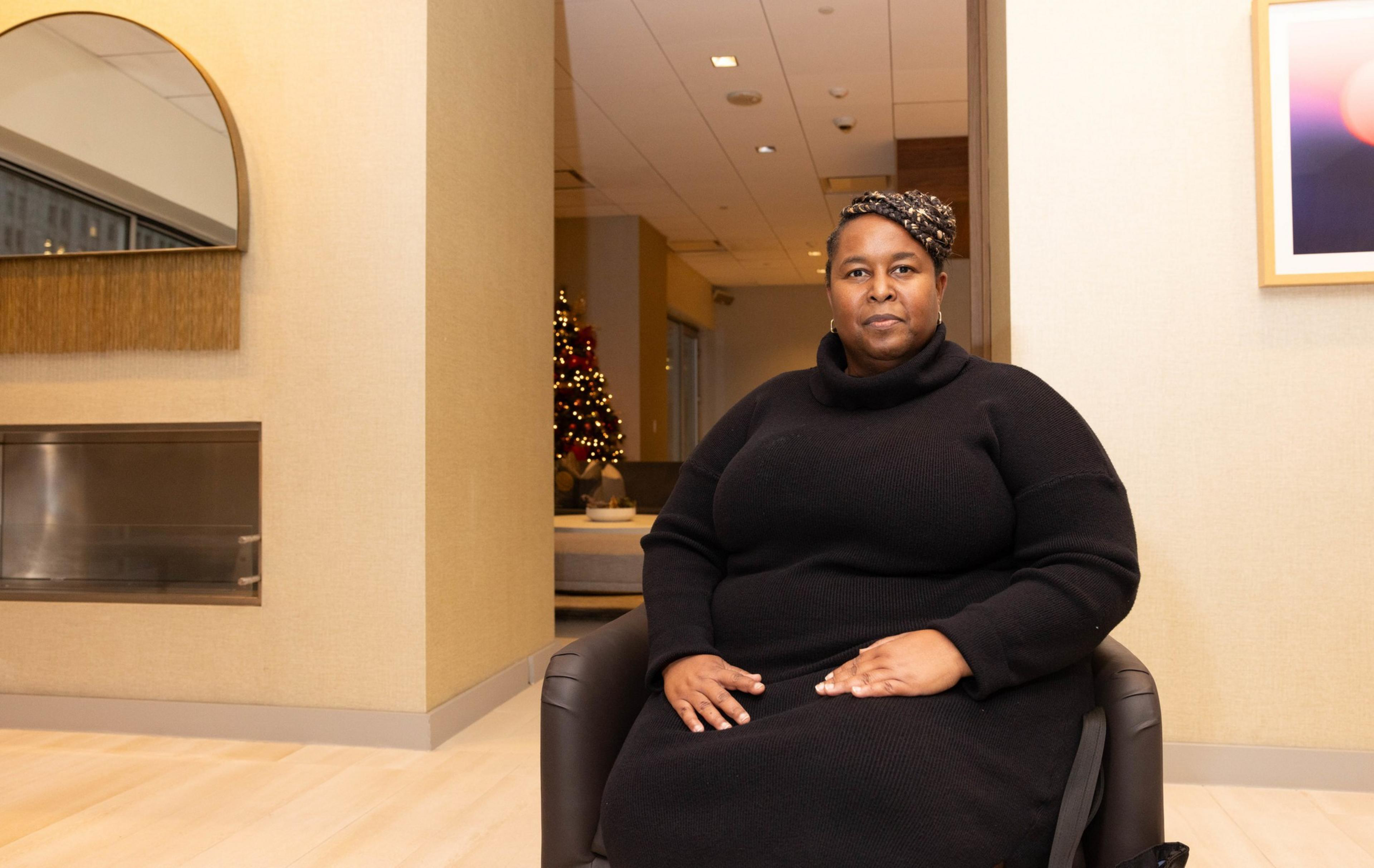 A person in a black dress sits on a chair with their hands on their lap. 
