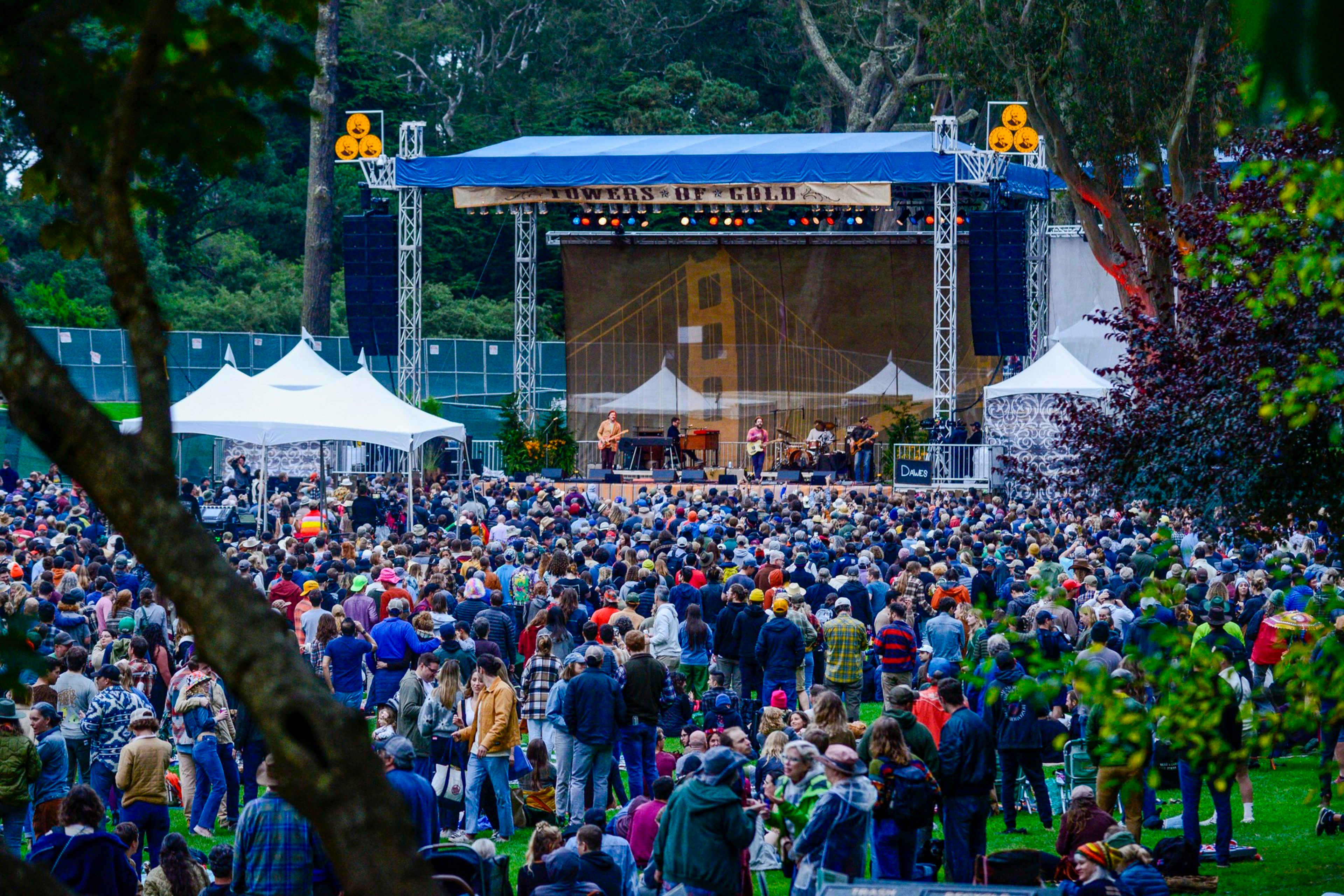 A band performs on a large outdoor stage with a crowd gathered on a grassy area. The backdrop features a bridge design, and trees surround the venue.