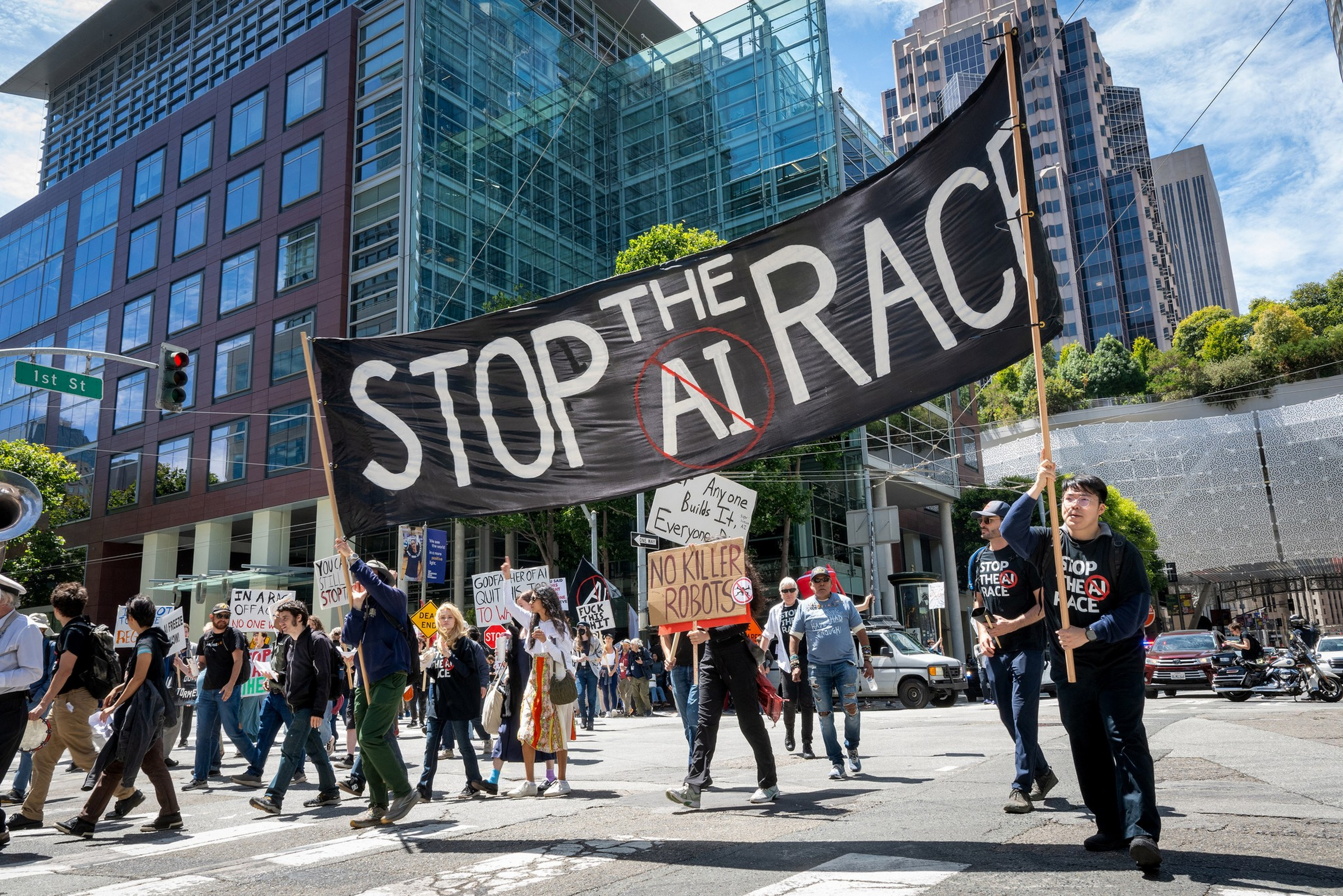 A crowd marches through a city street holding a large banner that reads "STOP THE AI RACE" and various signs opposing AI and killer robots.