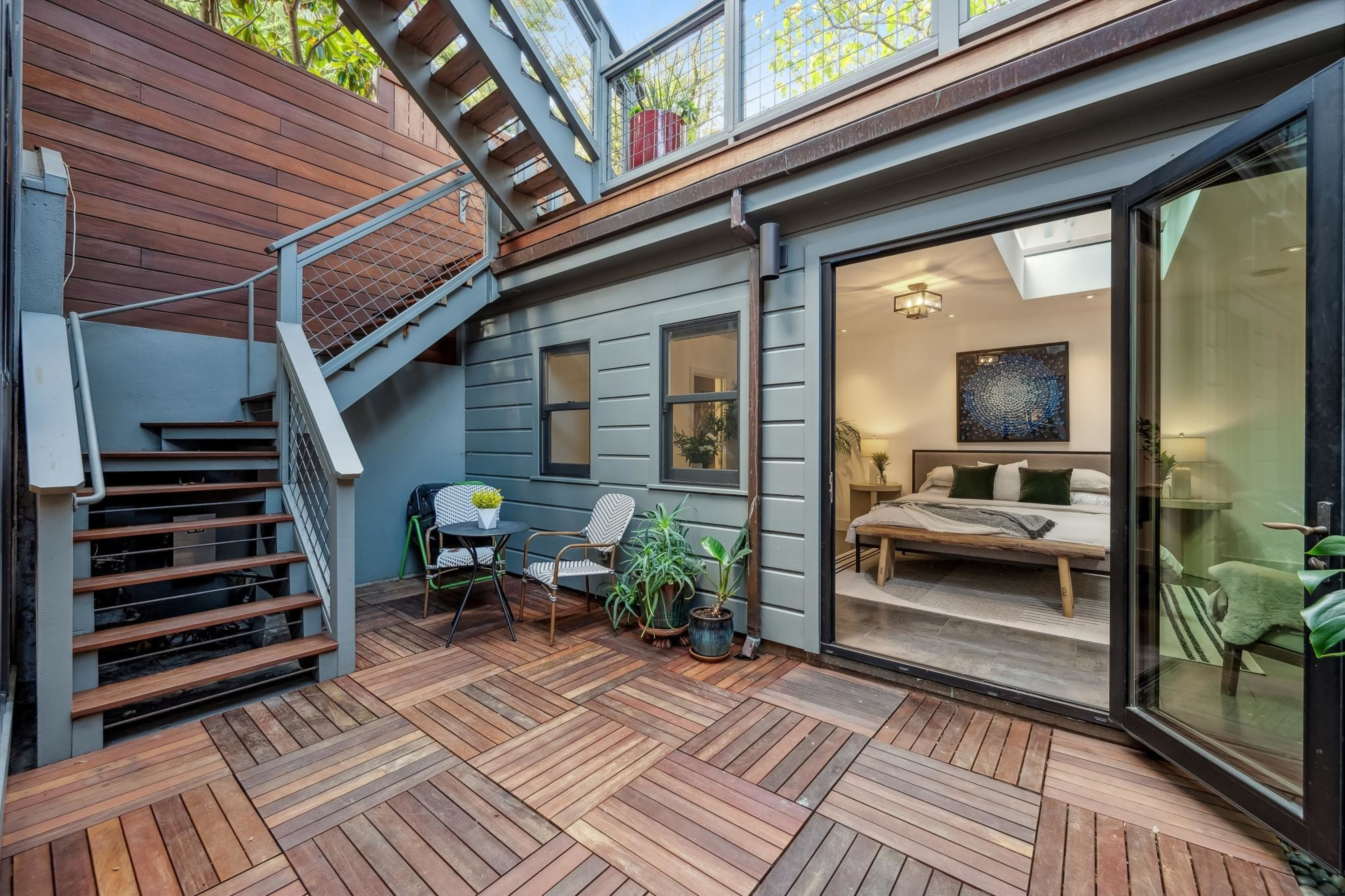 The image shows a cozy outdoor wooden patio with stairs leading up, two chairs, a small table, plants, and a view into a bedroom through glass doors.