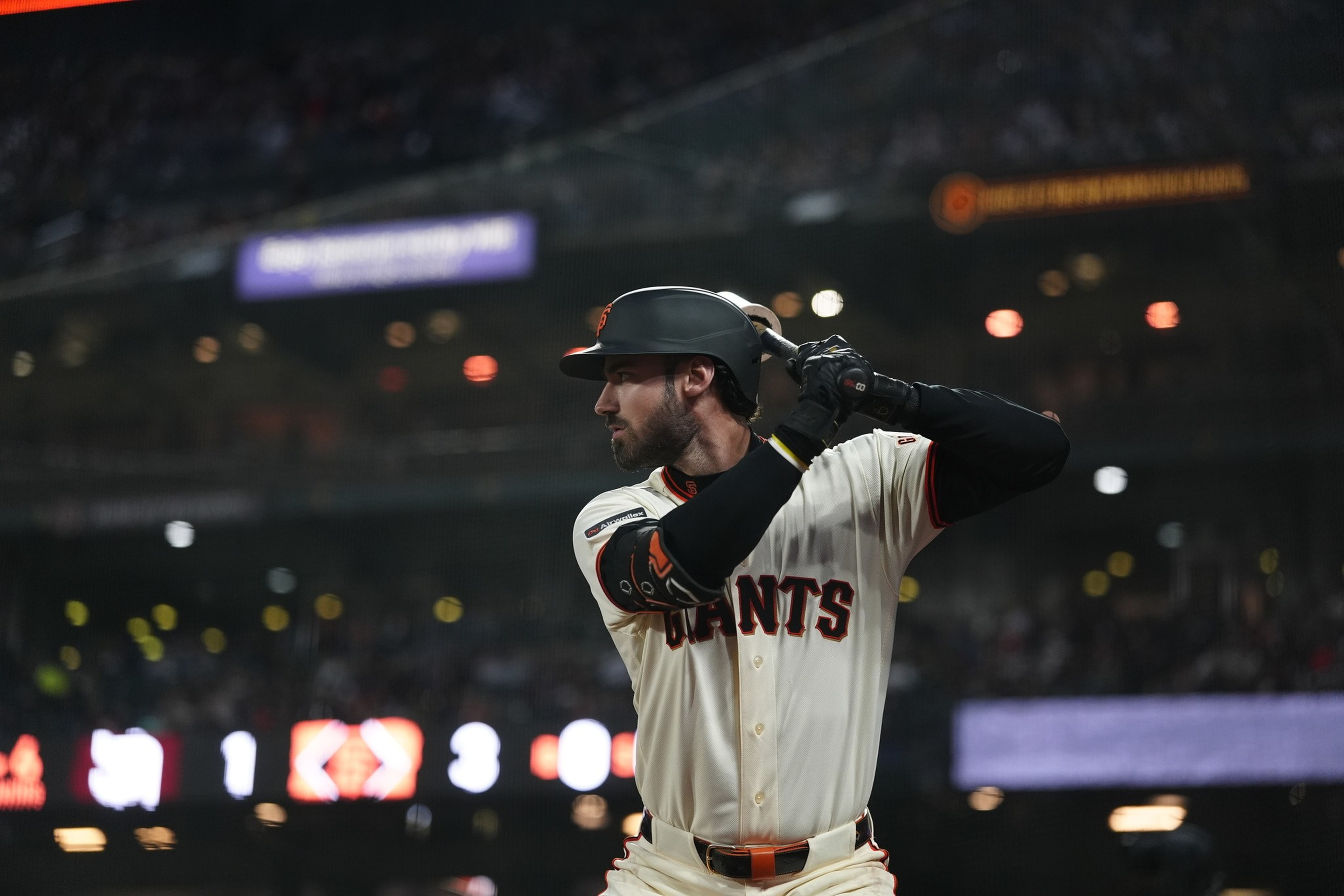 A baseball player from the Giants, wearing a helmet and batting gloves, stands ready to bat in a stadium under bright lights.