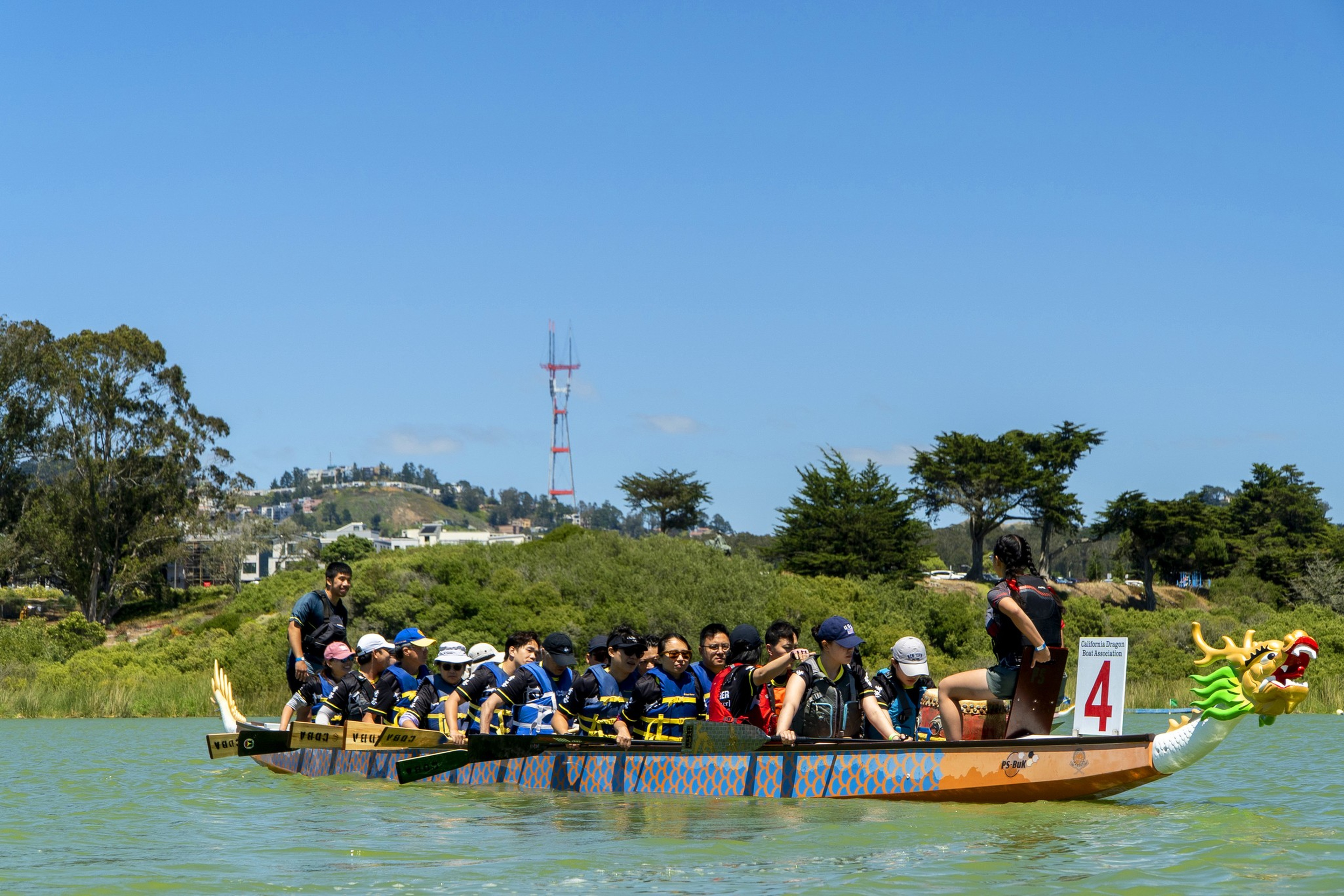 A group of people wearing life jackets row a dragon boat with a colorful dragon head and tail on a calm lake under a clear sky.
