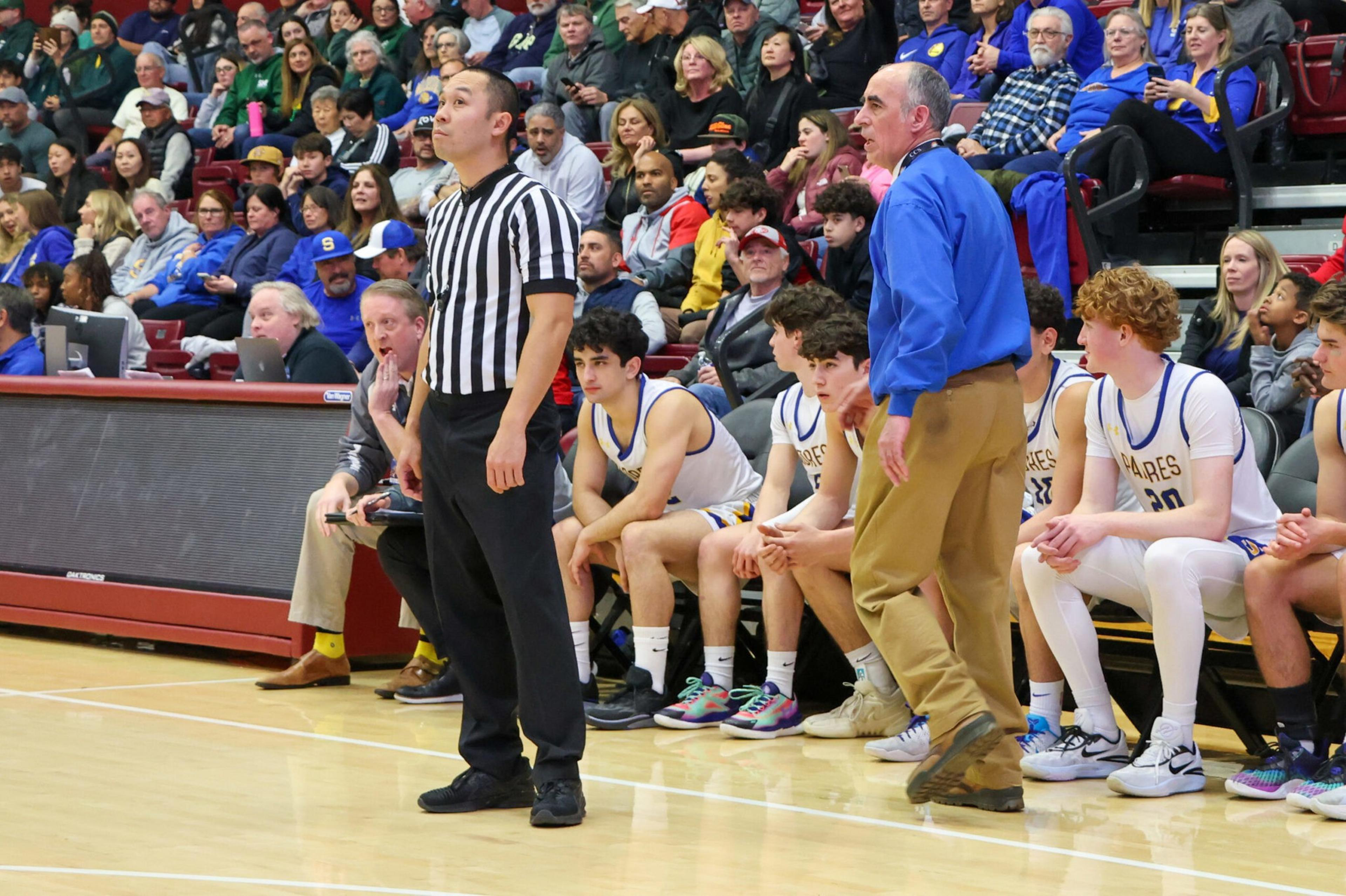A basketball referee stands near the bench watching the game, while players in white uniforms and a coach in blue focus intently on the court.