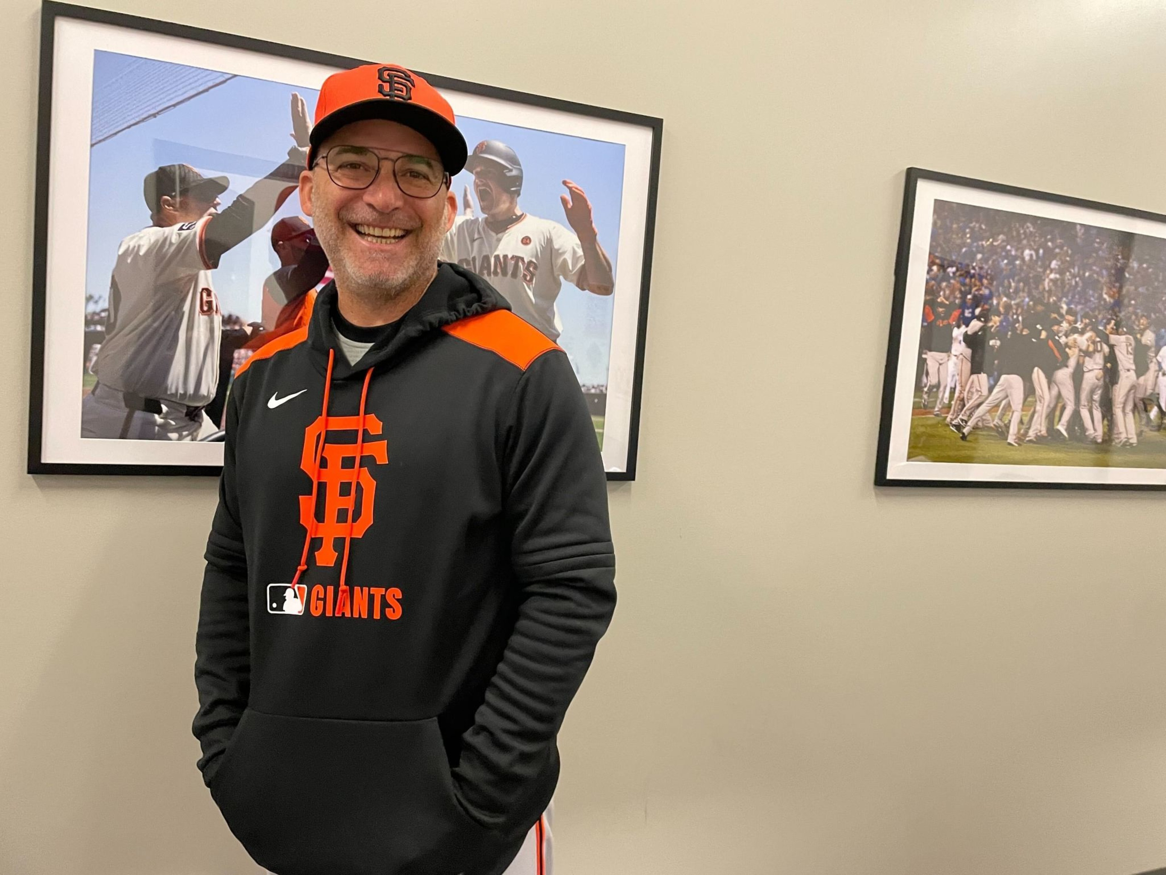 A smiling man in a San Francisco Giants hoodie and cap stands by two framed baseball photos on a beige wall.