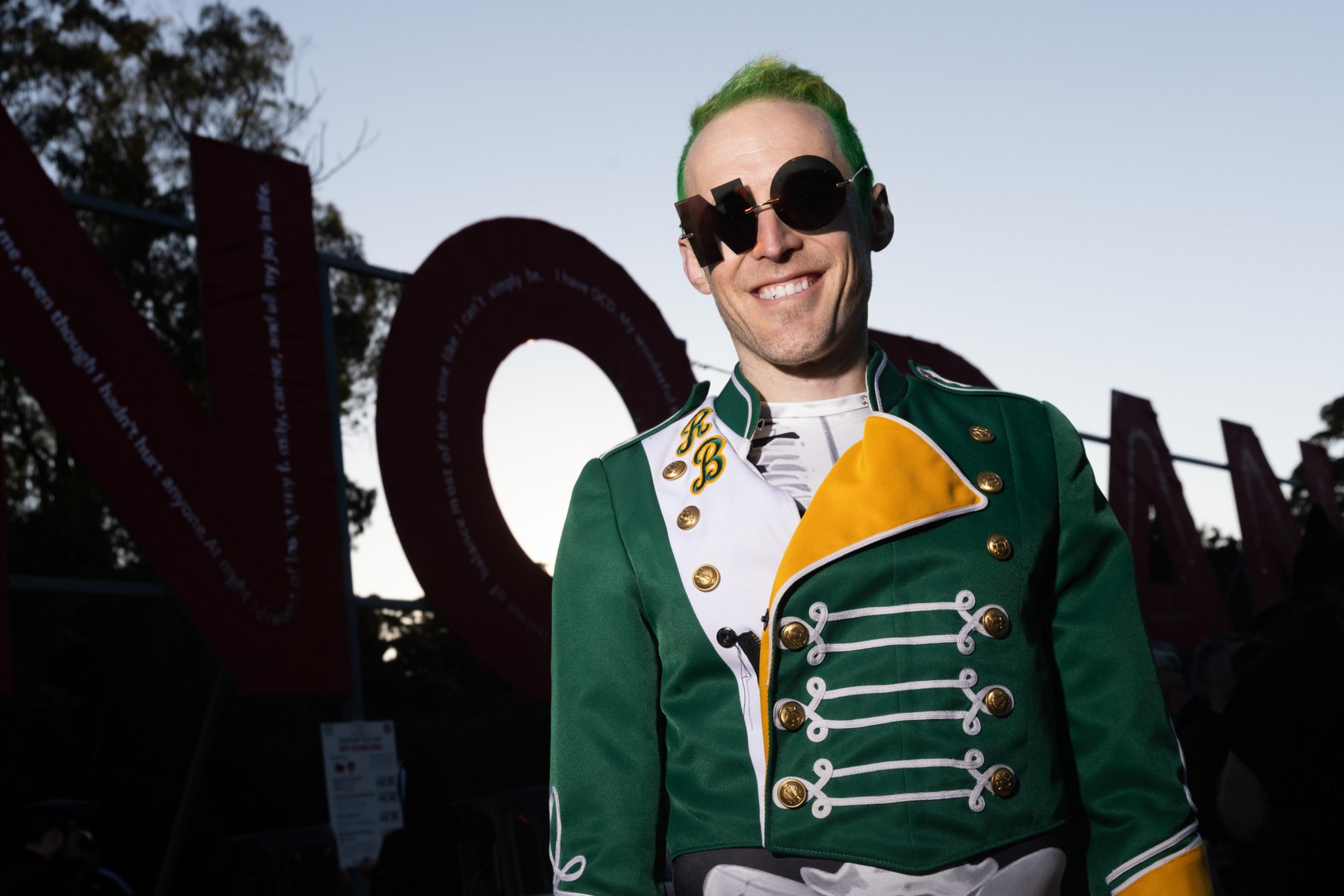 A person with green hair wears round black sunglasses and a green, white, and yellow marching band-style jacket while smiling outdoors.