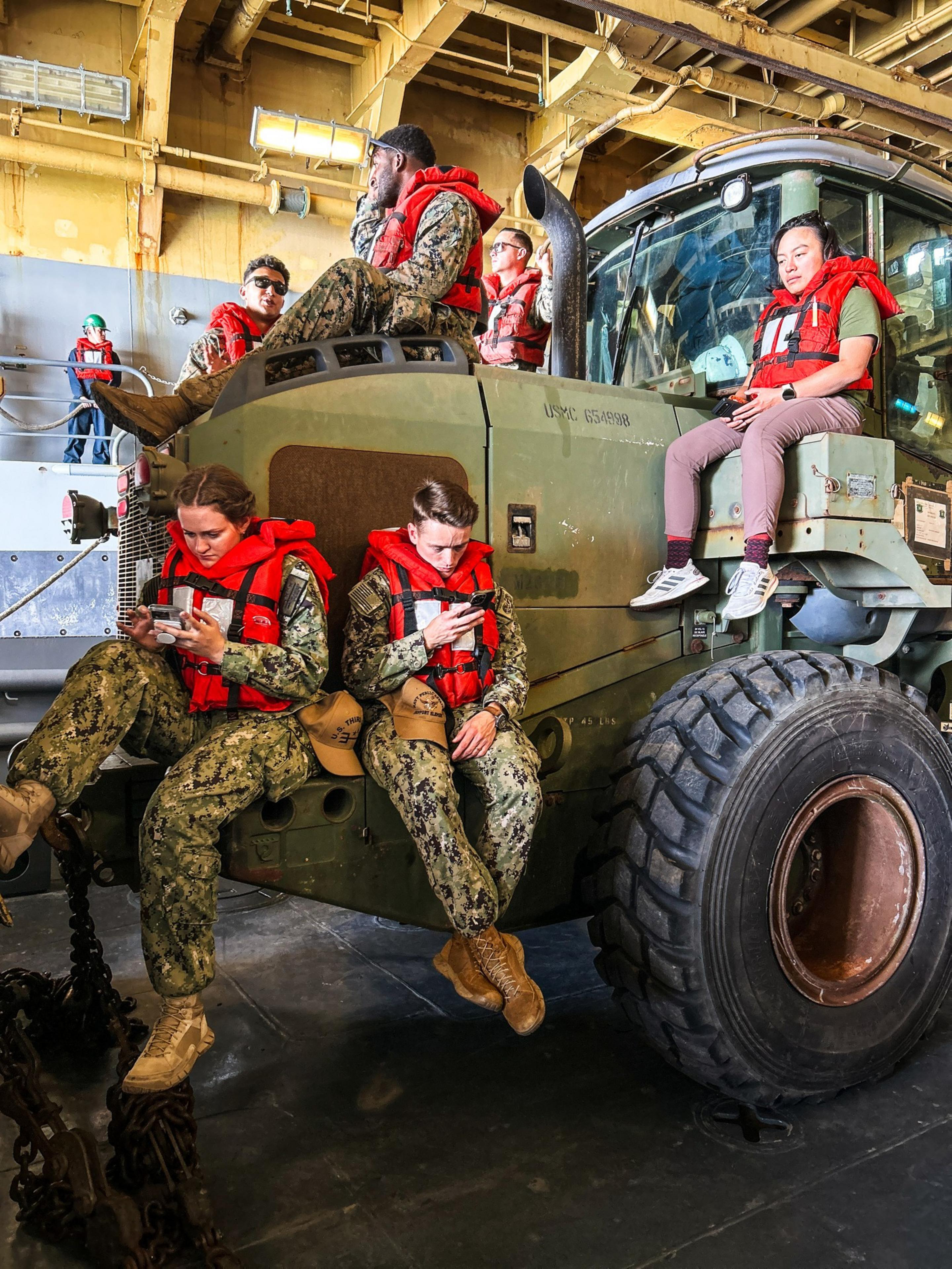 People in military uniforms and life vests are sitting and standing on a large vehicle inside an industrial setting, engaging with their phones.