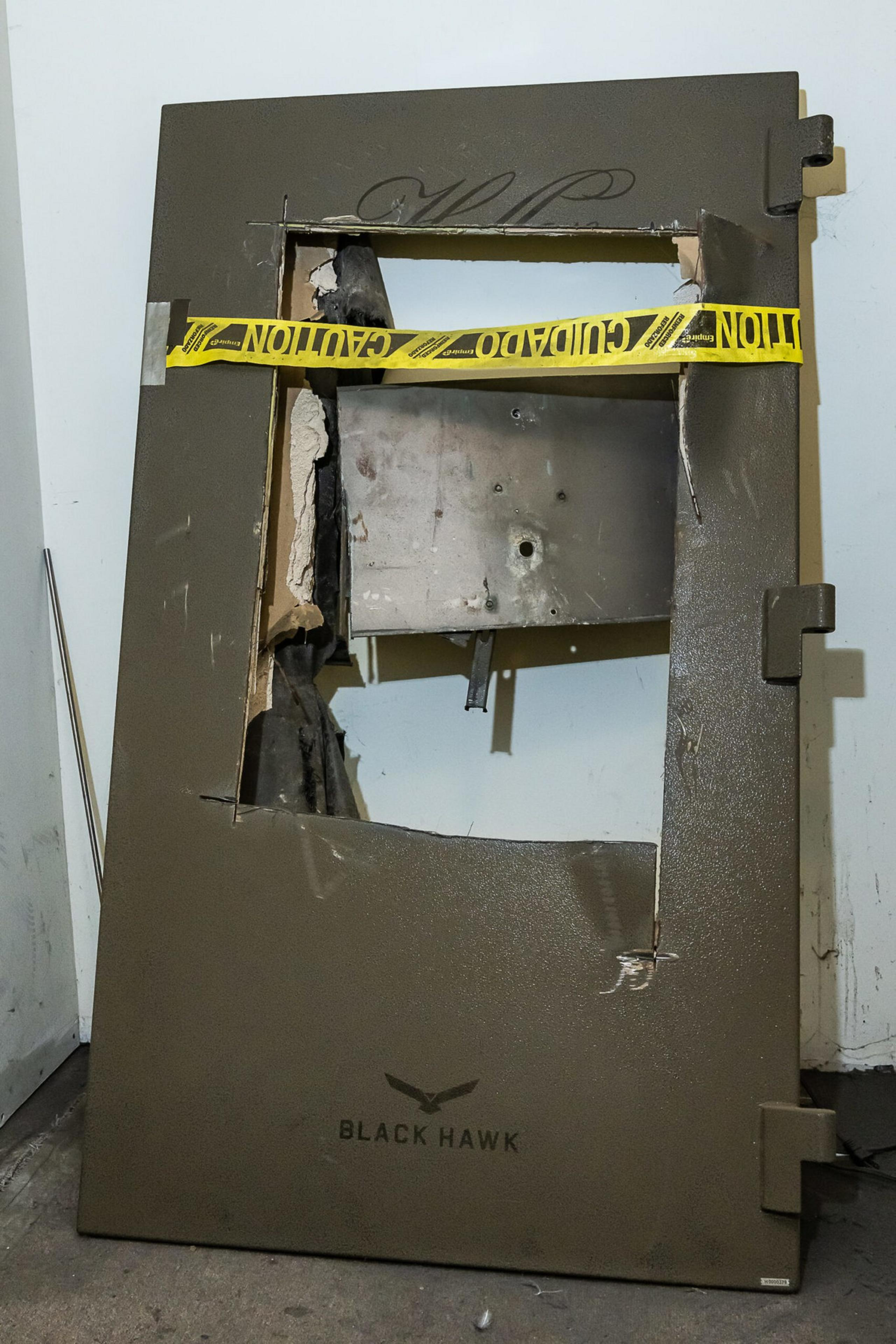 A large metal door with a jagged rectangular hole cut out is leaning against a wall, secured with yellow caution tape that reads "CAUTION CUIDADO."
