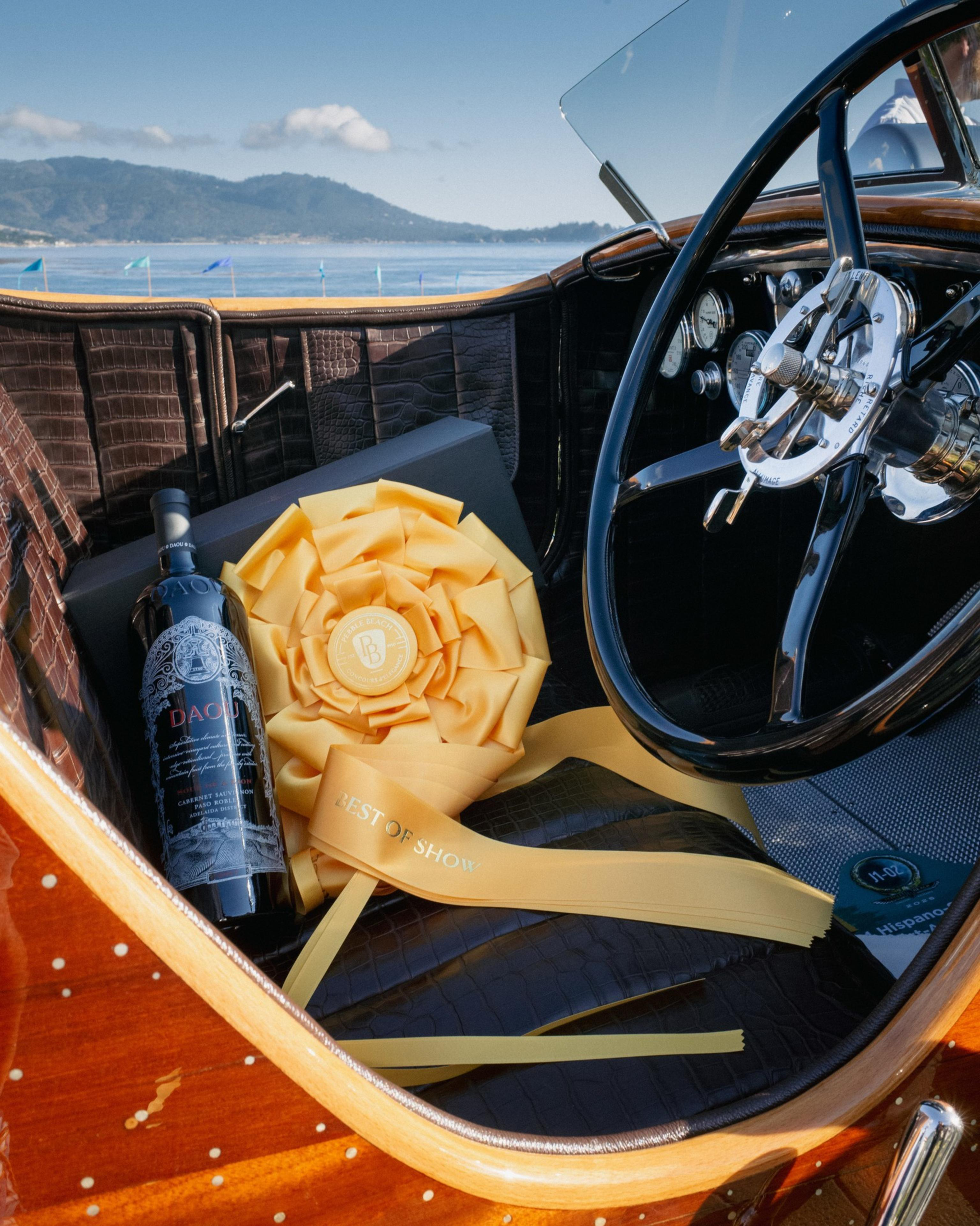 A leather-interior car seat holds a bottle of wine, a large yellow "Best of Show" ribbon, and a black box, with a steering wheel nearby.