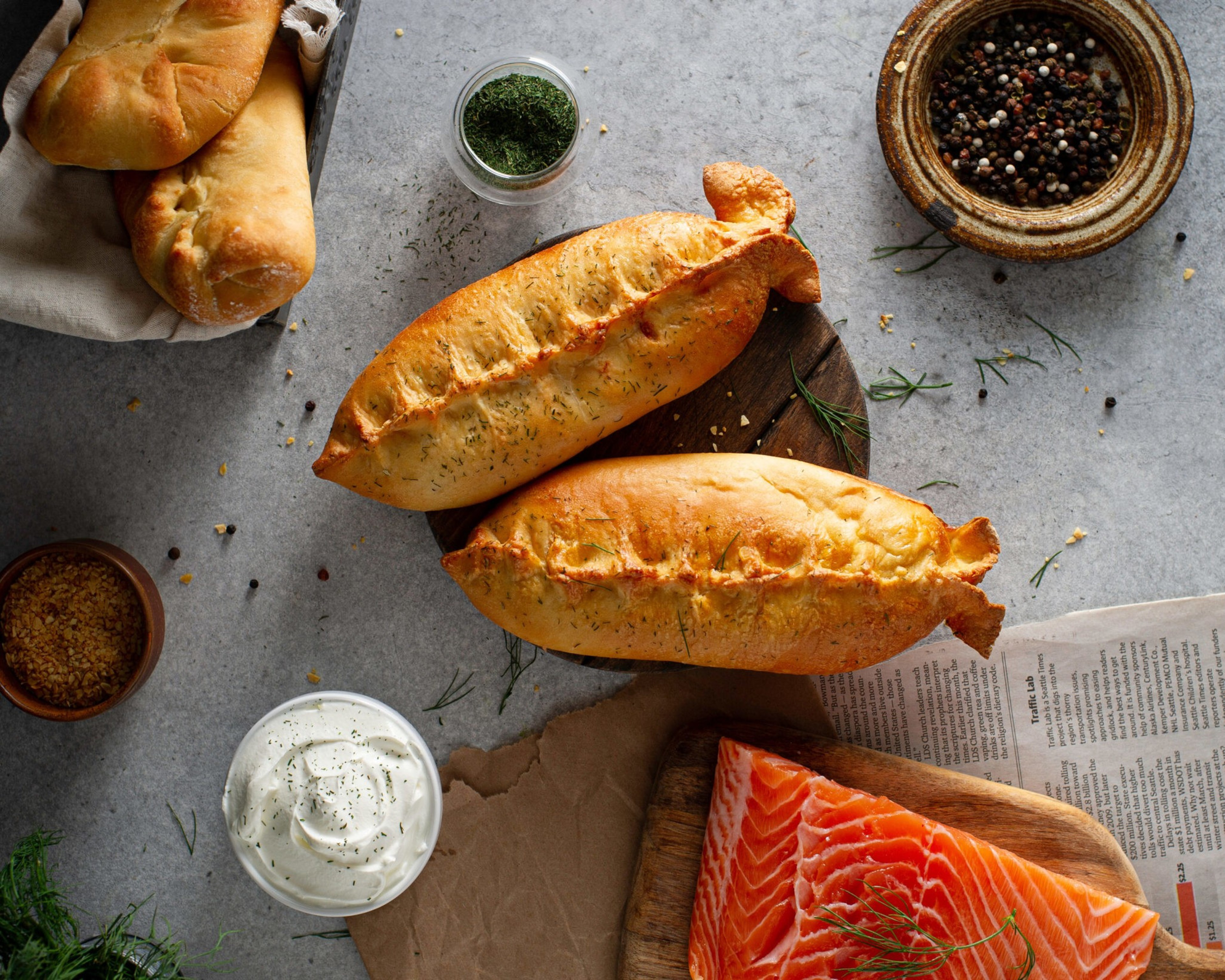 Two golden-brown baked pastries with herbs lie near a piece of raw salmon on a wooden board, surrounded by bowls of spices and creamy sauce.