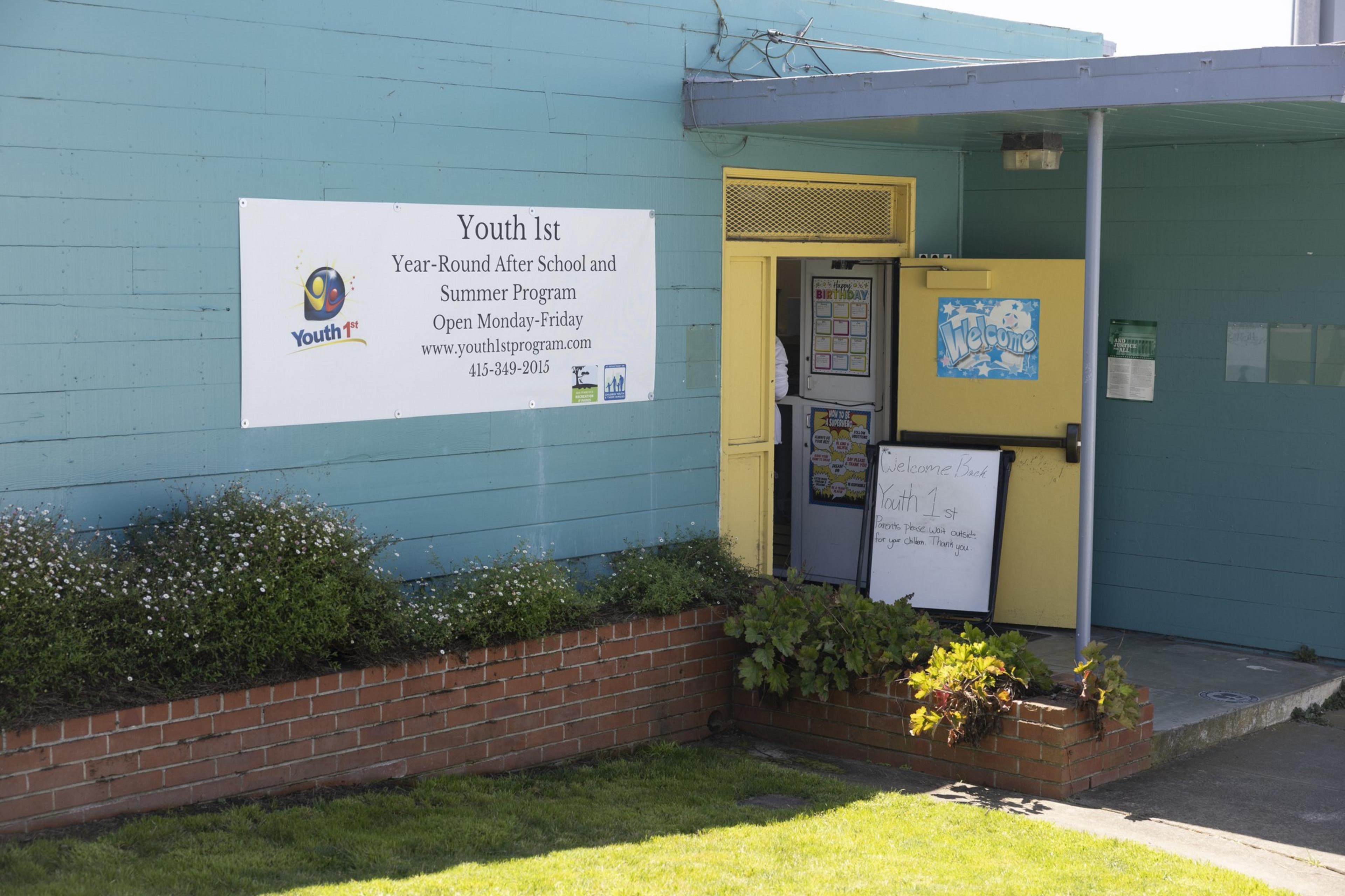 A teal building with a banner for "Youth 1st Year-Round After School and Summer Program" and an open yellow door displaying welcome signs and a whiteboard.