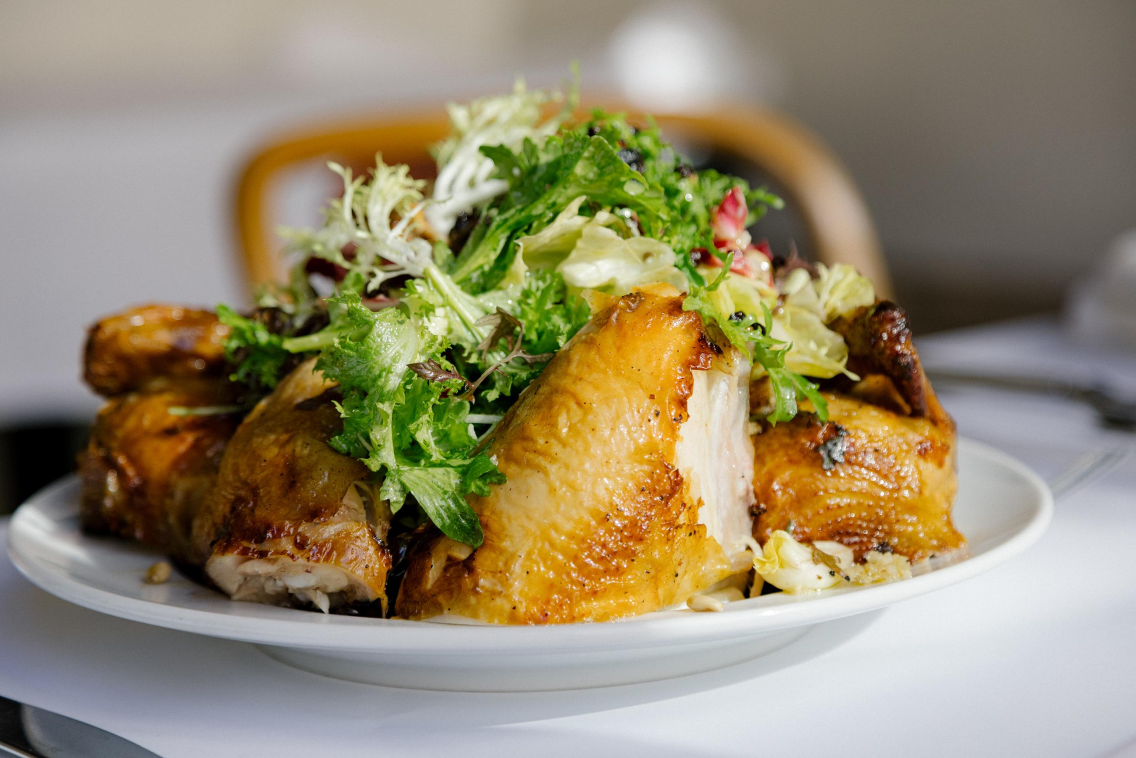 A tight detail photo of Chicken for two roasted in the wood-fired oven served with a warm bread salad with scallions, garlic, bitter greens, dried currant and pine nuts on a white table cloth.