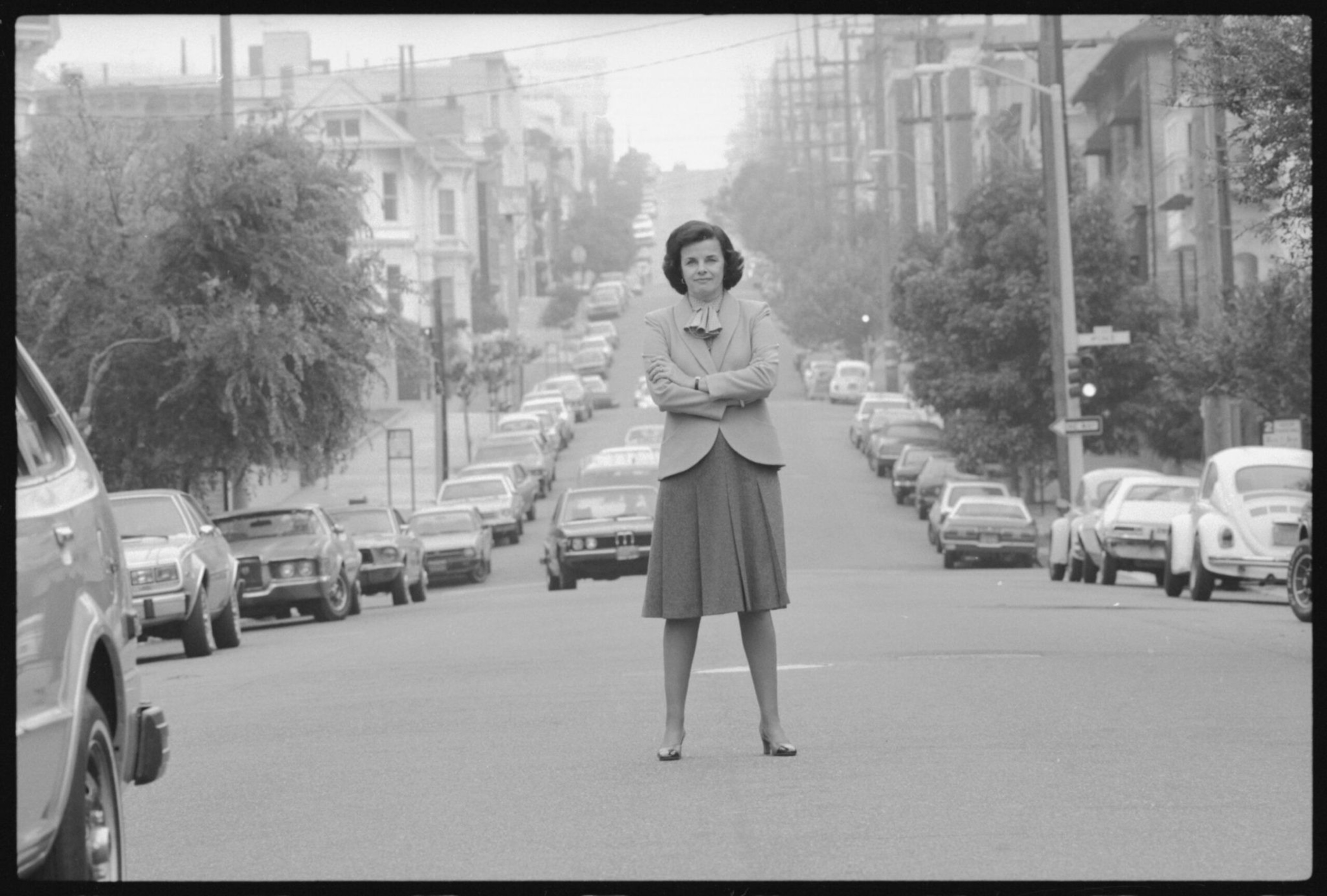 A woman in a skirt and blazer stands confidently in the middle of a street lined with parked cars and houses on both sides.