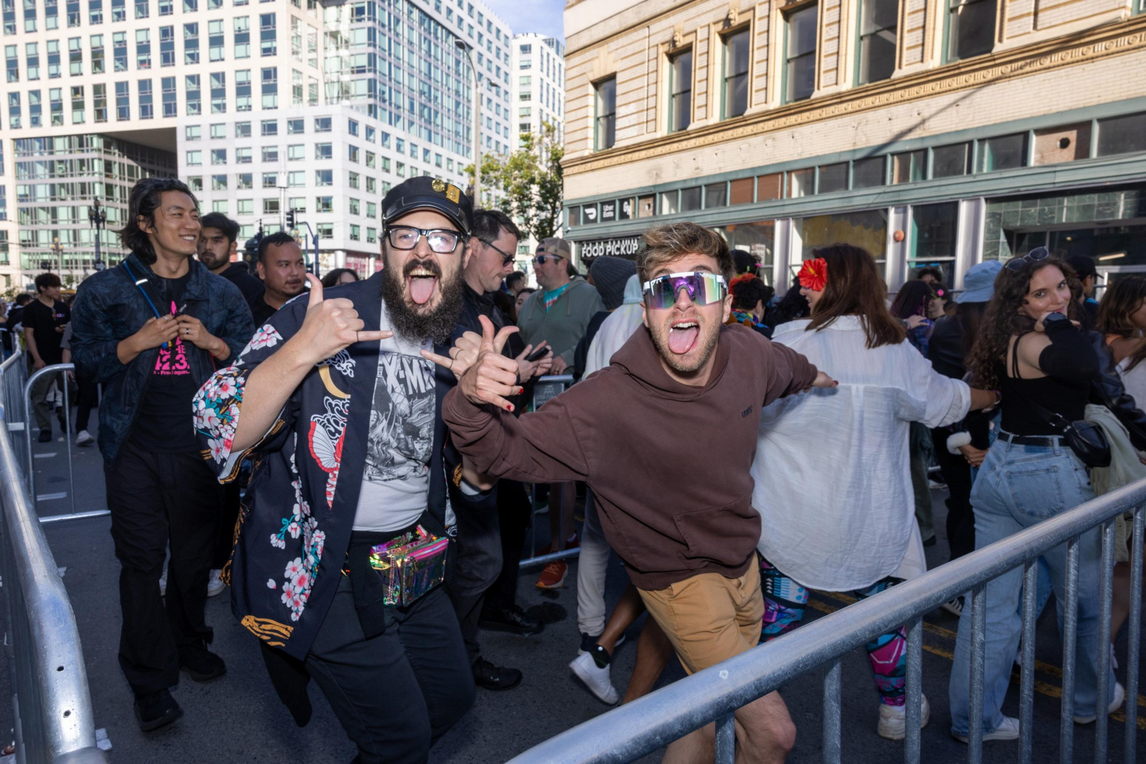 A lively group of people is gathered outside an urban setting with tall buildings. Two men in the foreground are posing energetically with tongues out and making hand gestures.