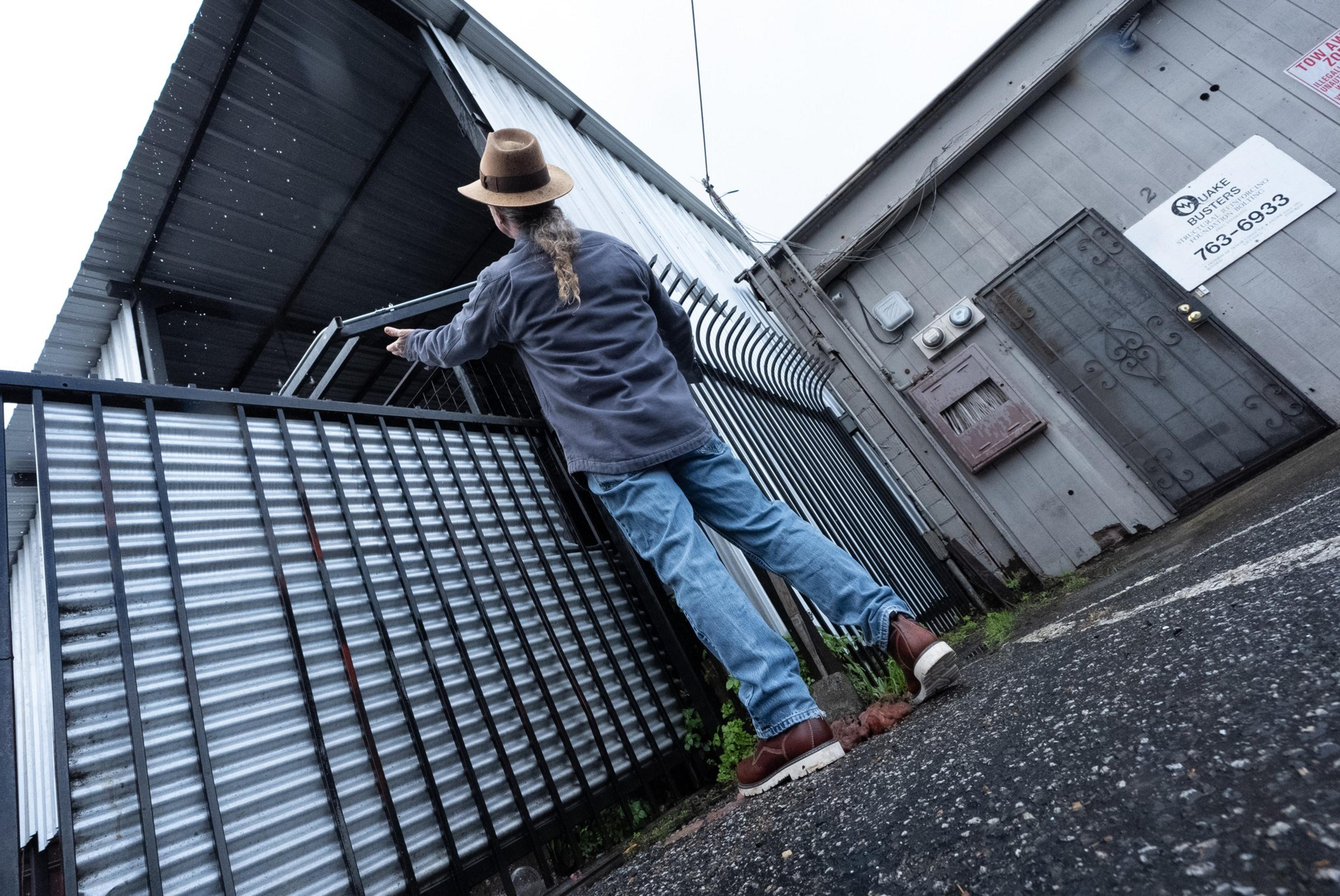 A person wearing a hat and denim jeans stands on wet ground, reaching over a corrugated metal fence beside a gray industrial building with a &quot;Quake Busters&quot; sign.
