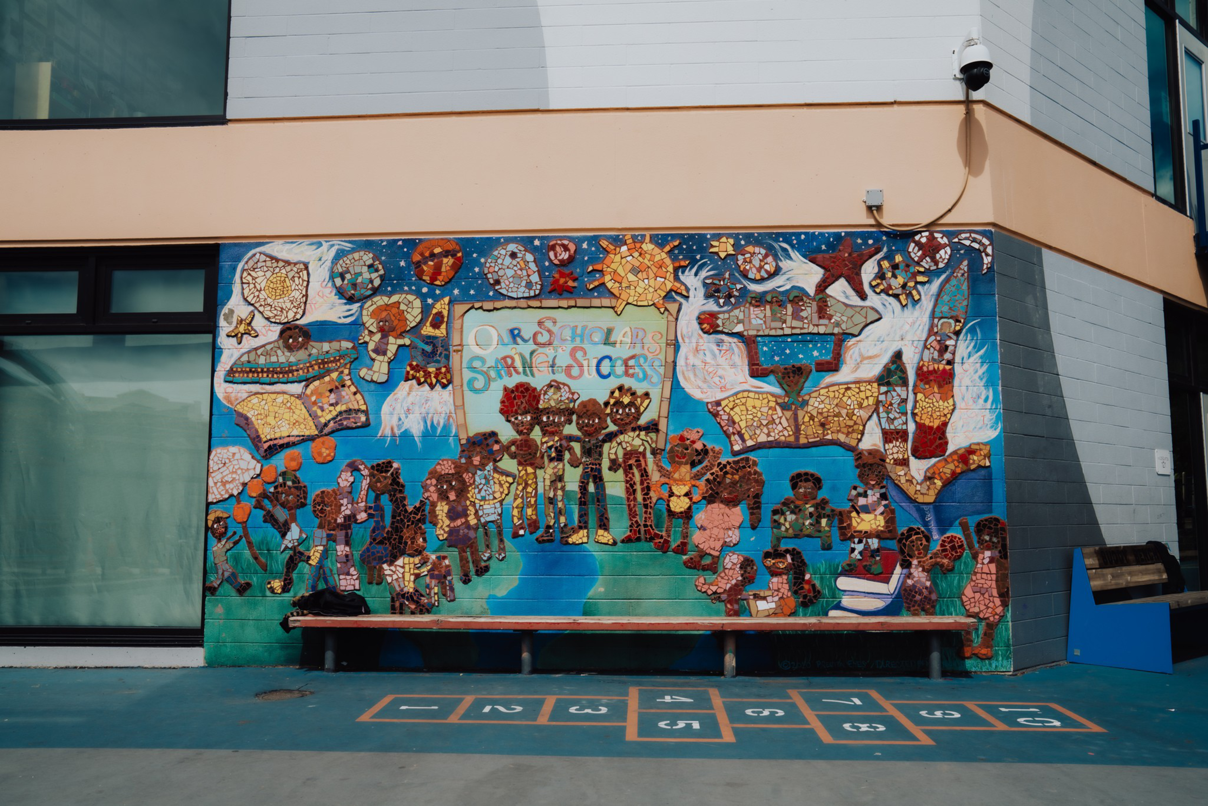 A colorful mosaic mural on a school wall features children, books, stars, and the phrase “Our Scholars Our Minds Our Success” with a hopscotch game in front.