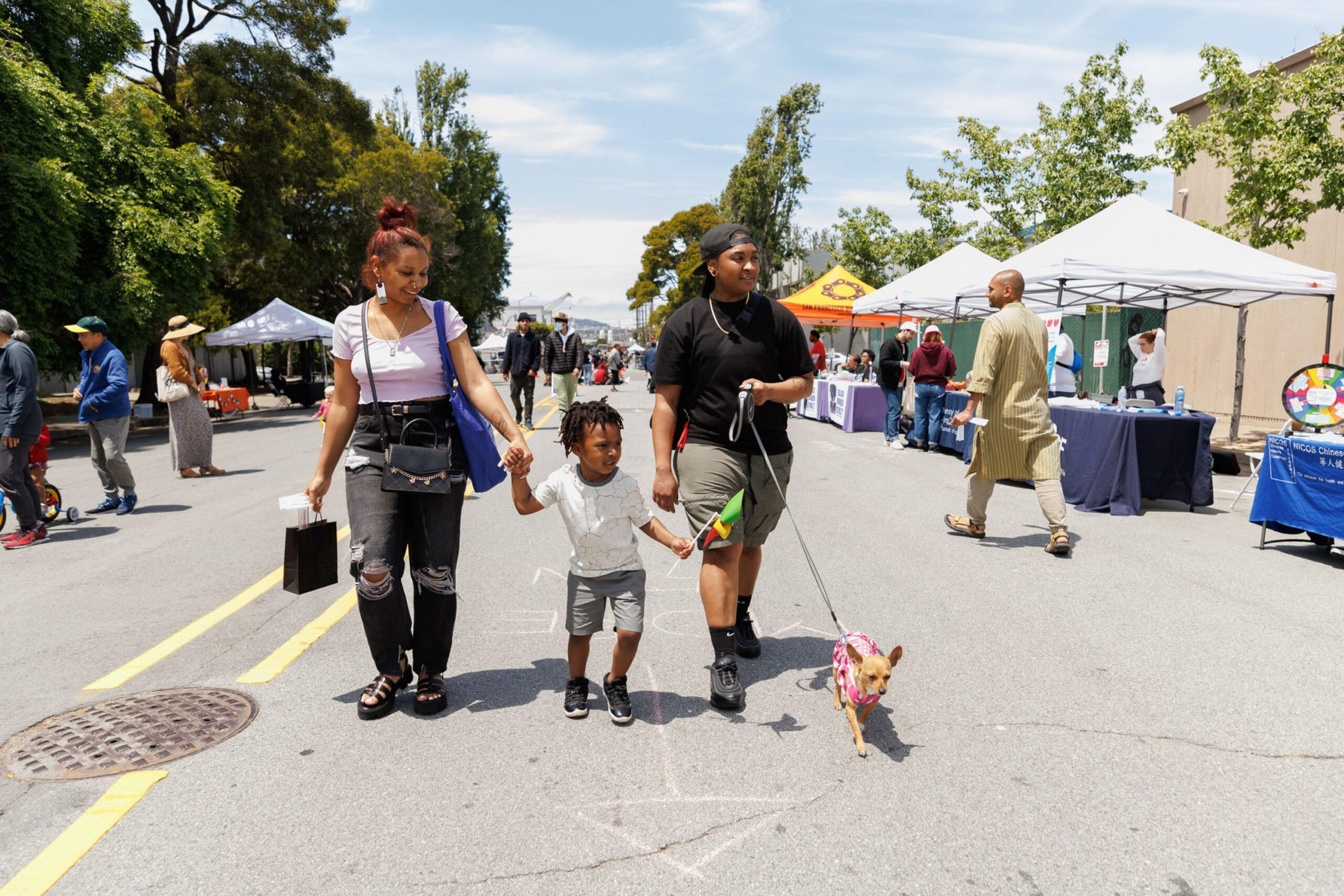 Two adults and a child walk a dog at a sunny street fair, past tents and other attendees.