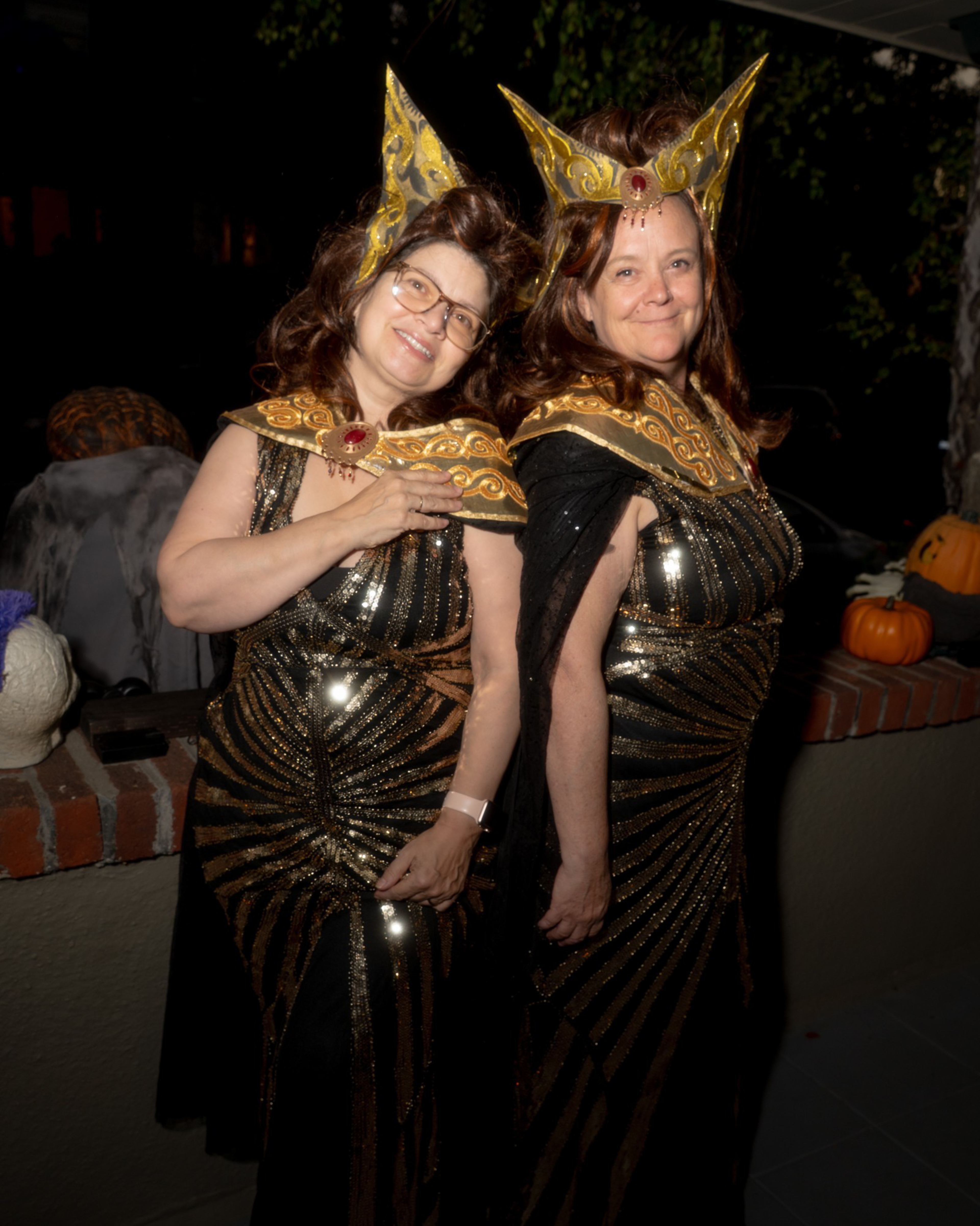 Two women wear matching black and gold shimmering gowns with ornate golden collars and tall, pointed golden headpieces, smiling and posing together.