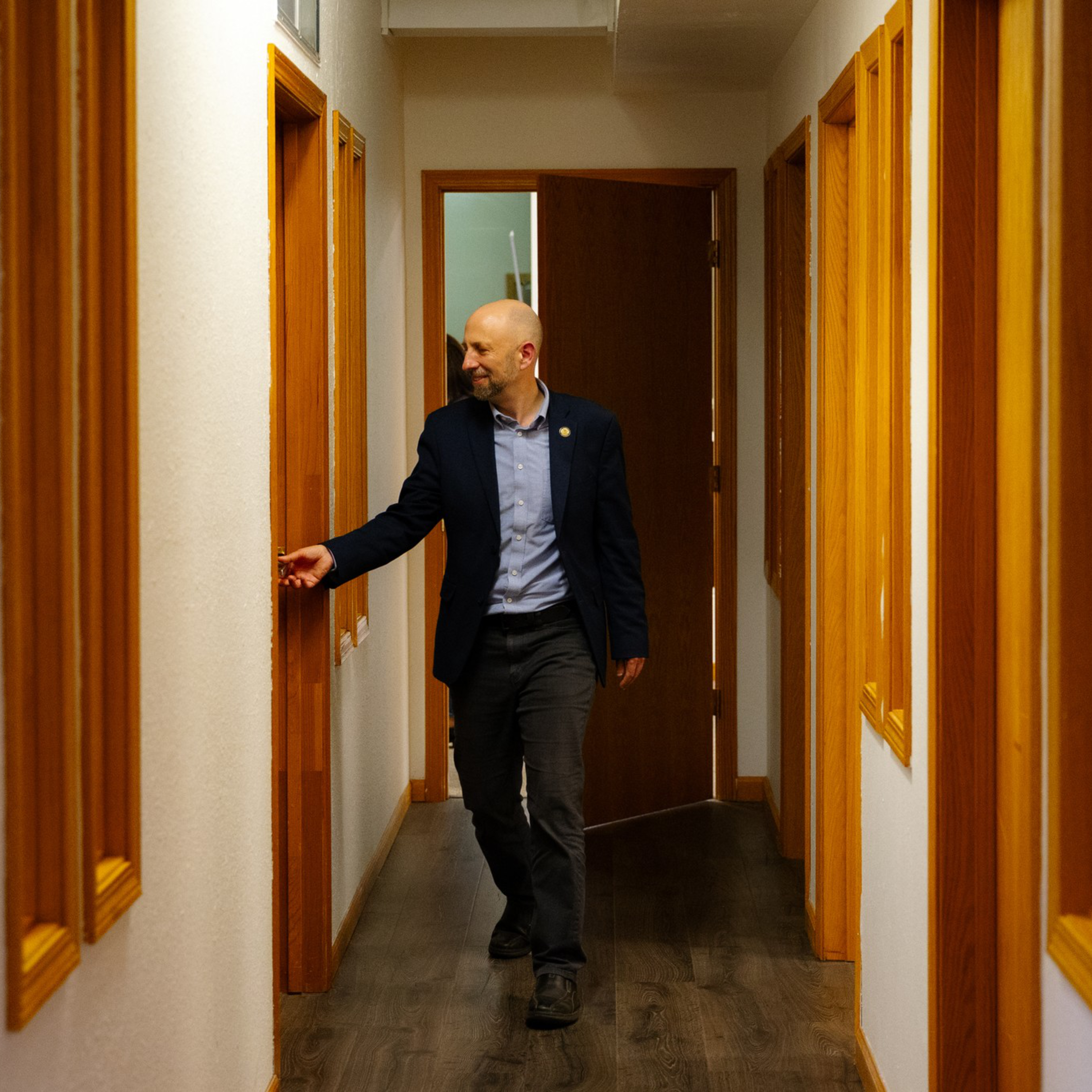 A bald man in a dark blazer and light blue shirt walks down a hallway with wooden-framed windows, reaching out to touch a light switch.