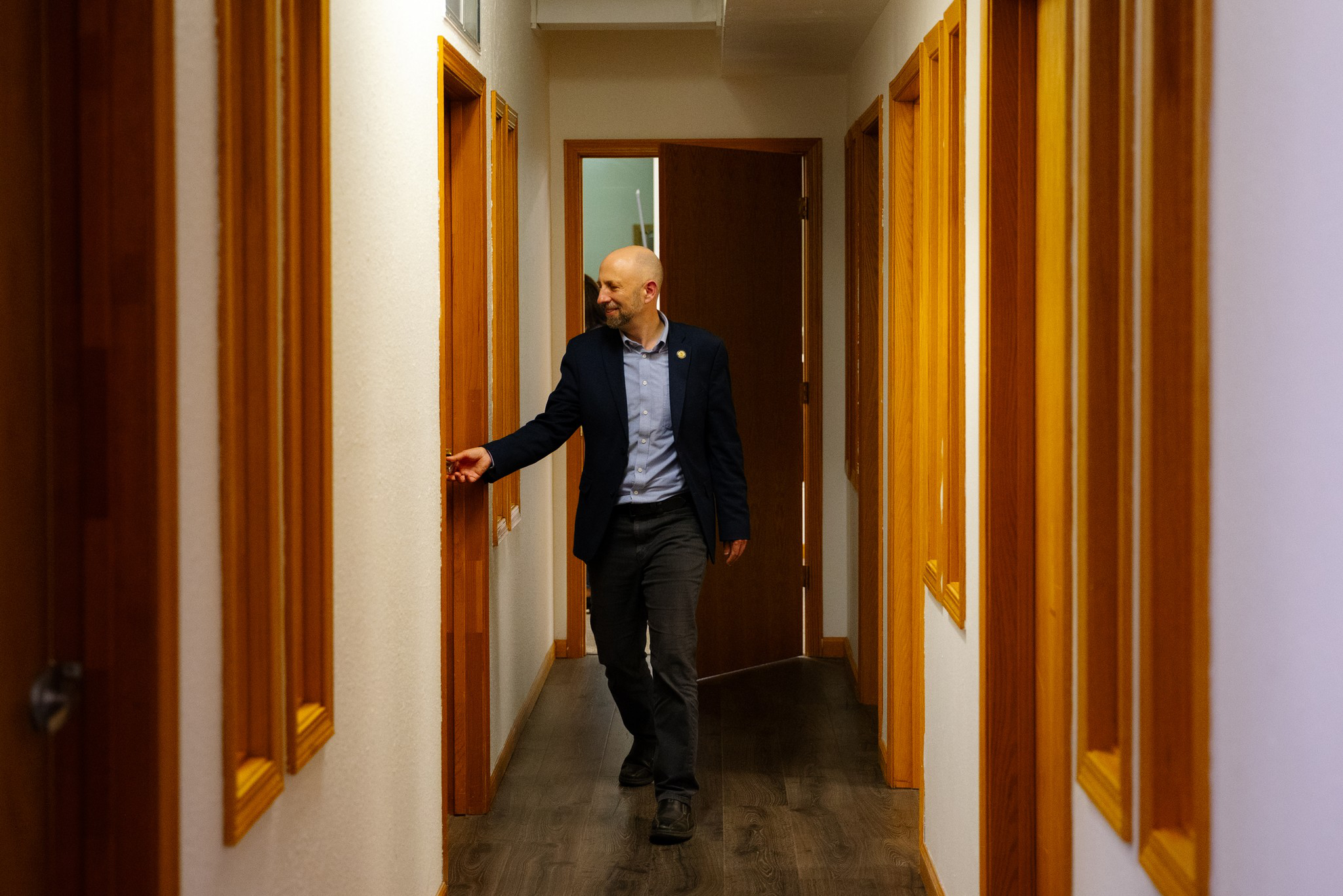 A bald man in a dark blazer and light blue shirt walks down a hallway with wooden-framed windows, reaching out to touch a light switch.