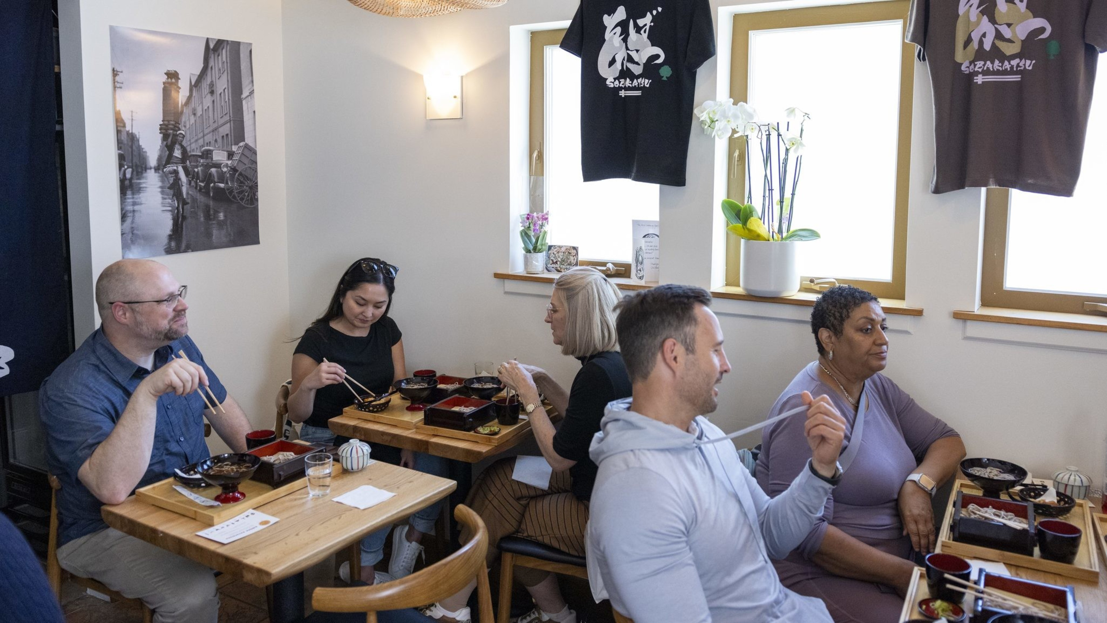 Five people are sitting at wooden tables in a cozy restaurant, eating with chopsticks. The room has a warm atmosphere with hanging shirts and a wall photo.