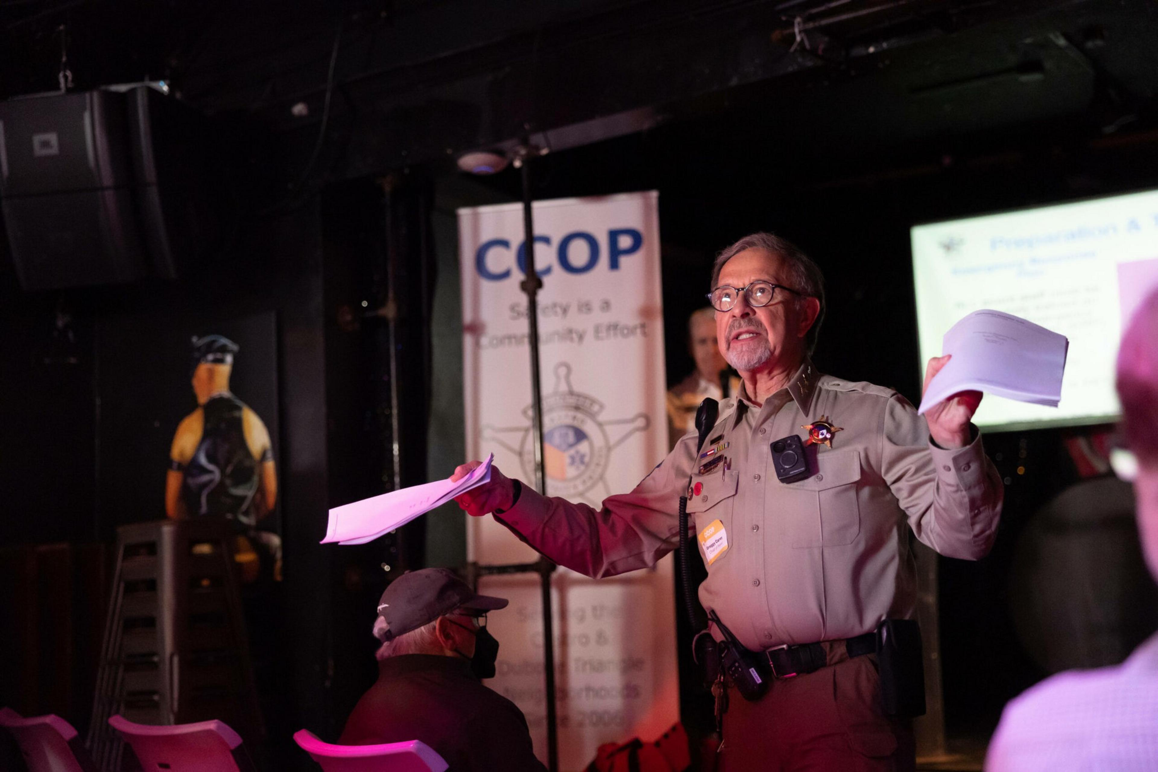 A uniformed officer with glasses holds papers and speaks to an audience in a dimly lit room with a "CCOP" safety banner behind him.