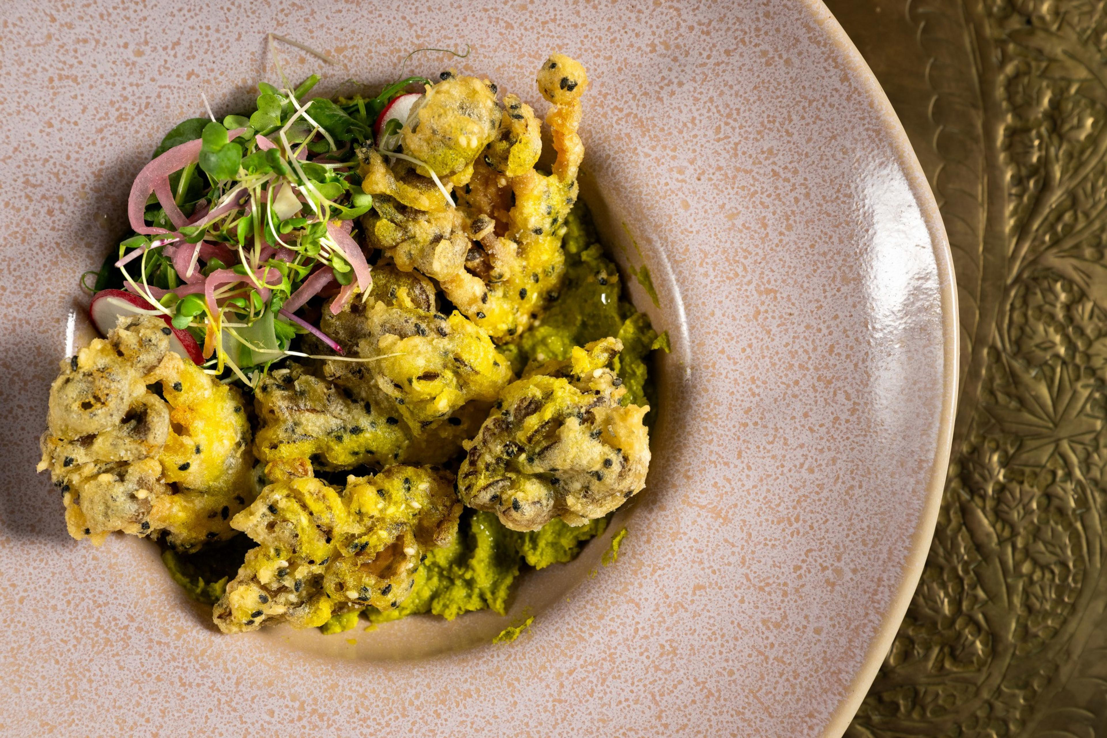 The image shows crispy, golden-brown fried fritters on a plate, accompanied by vibrant green sauce and garnished with fresh herbs and pink pickled onions.
