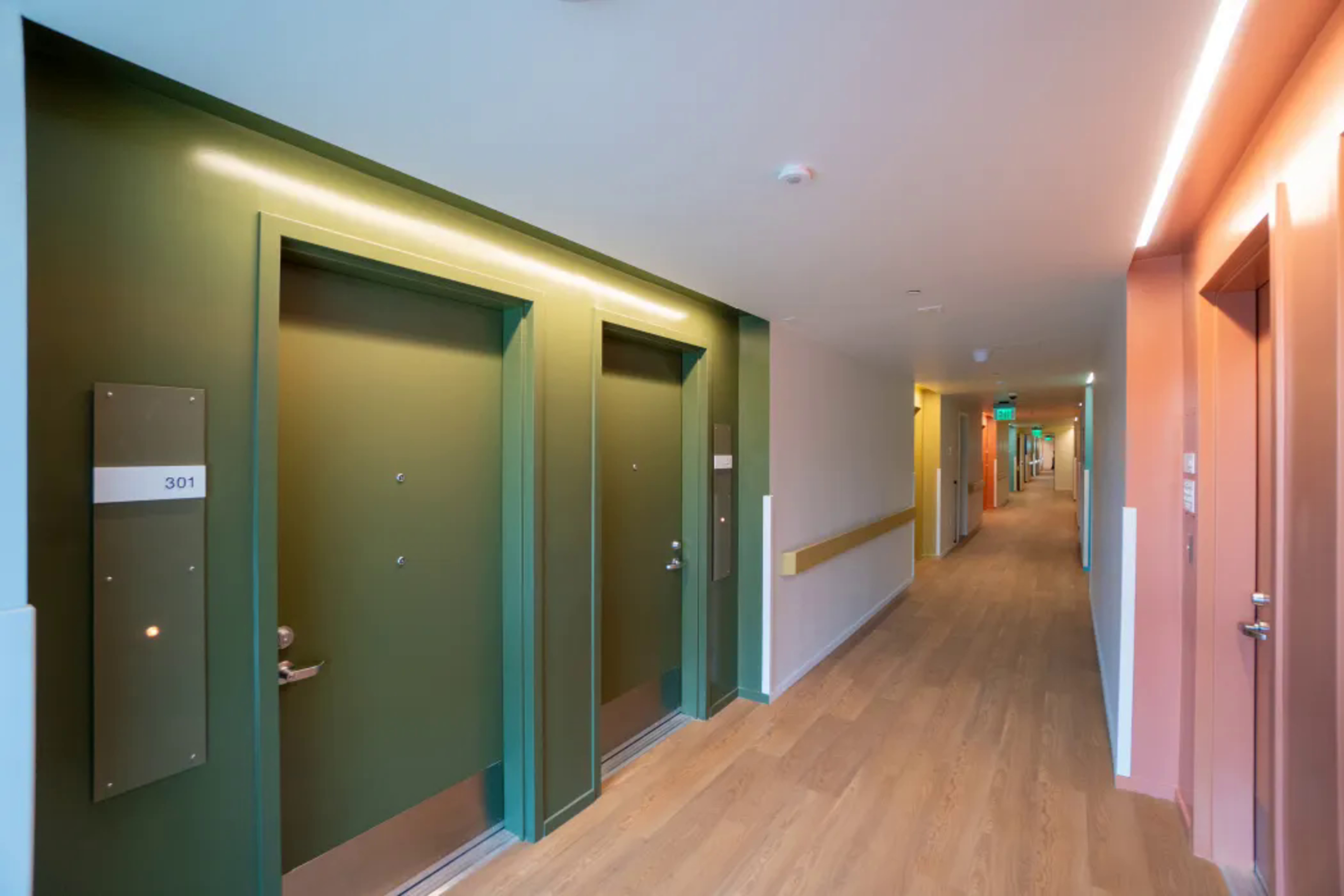 A modern hallway with wooden flooring features green doors on the left and pink doors on the right, with soft overhead lighting and a handrail on one wall.