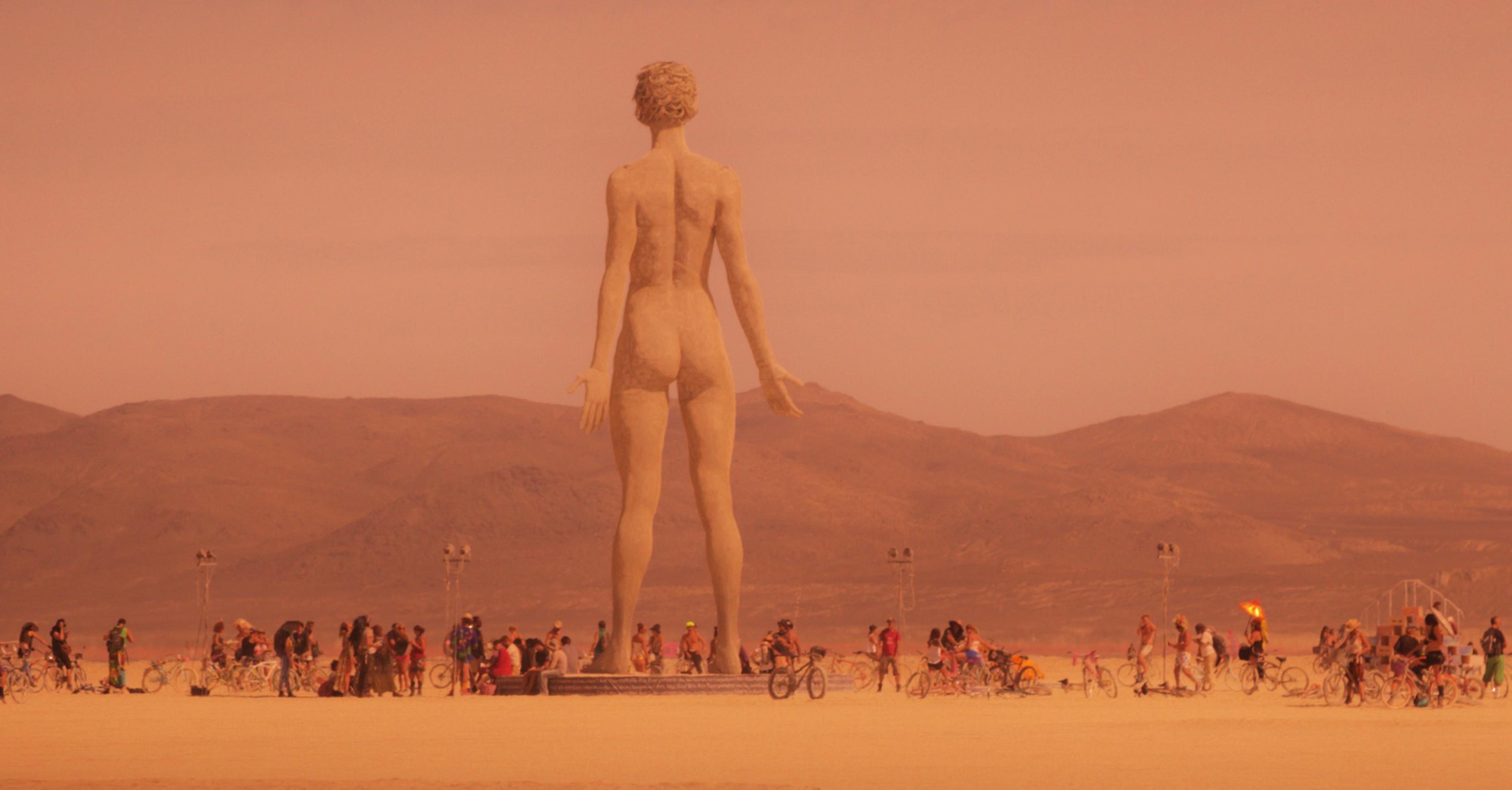 A giant sculpture of a standing human figure dominates a desert landscape, surrounded by people and bicycles, with mountains in the background under a reddish sky.