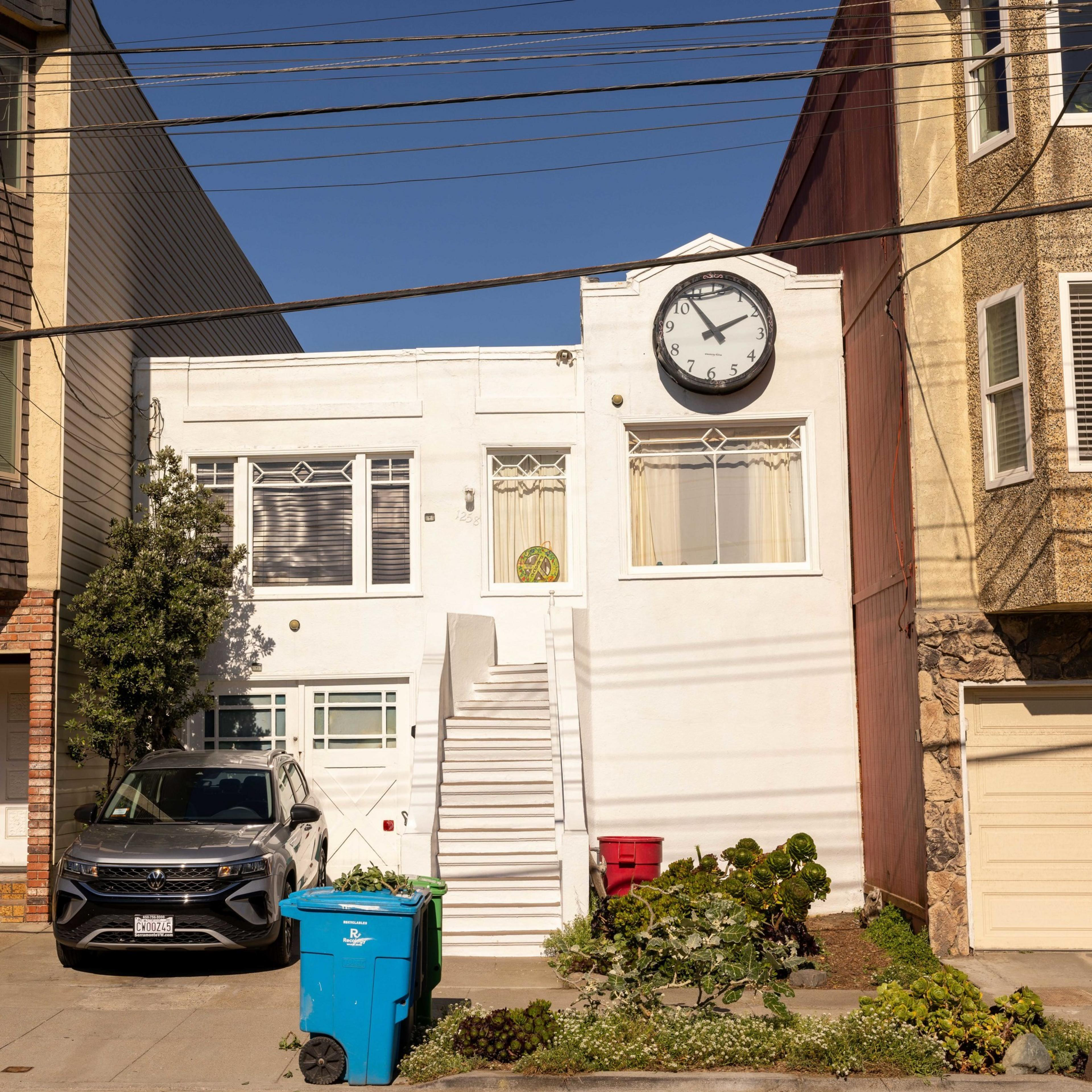 A white house with a large clock above the door, flanked by plants and recycling bins on the sidewalk, and a parked car to the left.
