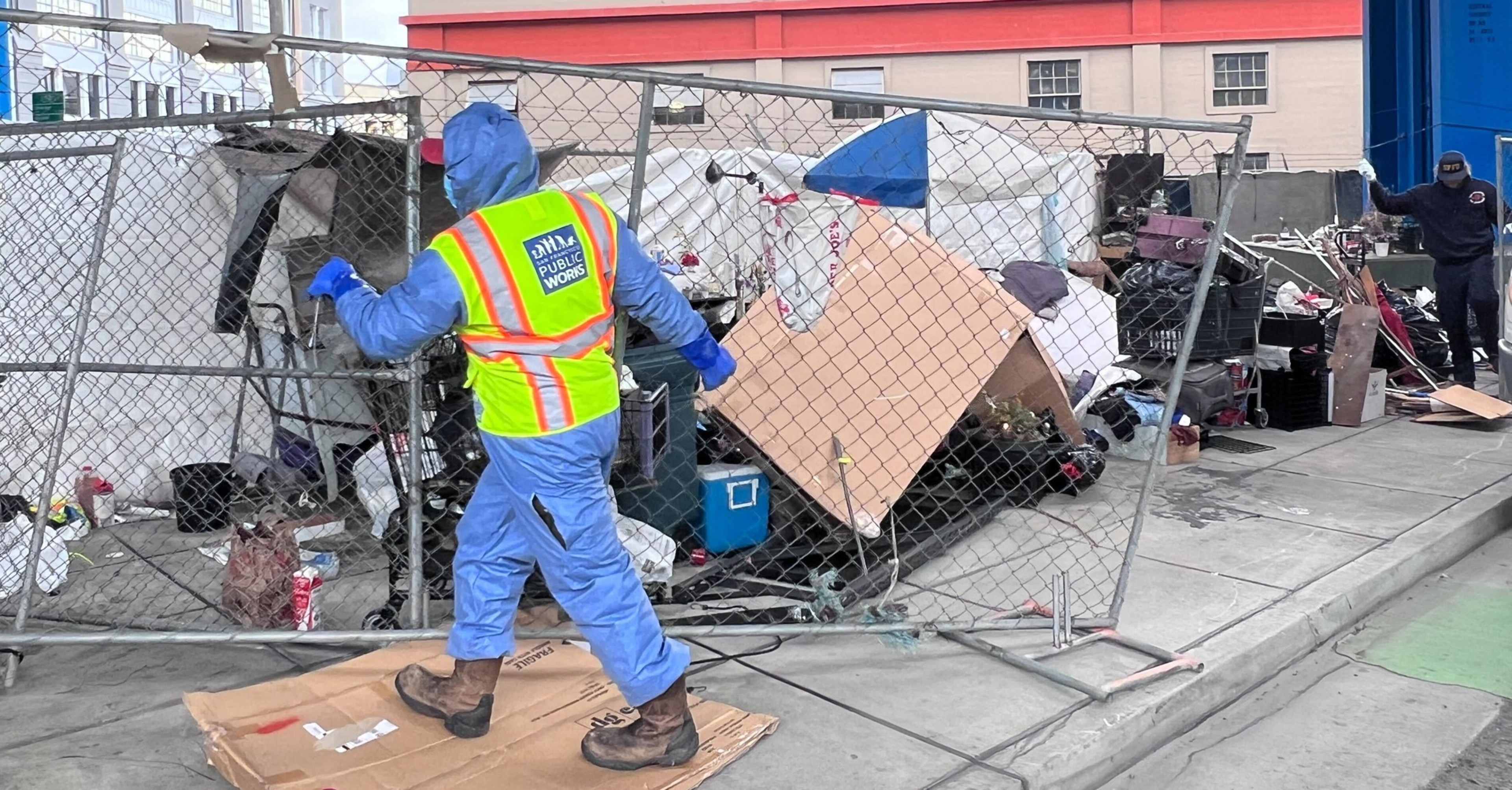 A worker in blue protective gear and a neon vest labeled "Public Works" removes a metal fence in front of a cluttered homeless encampment.